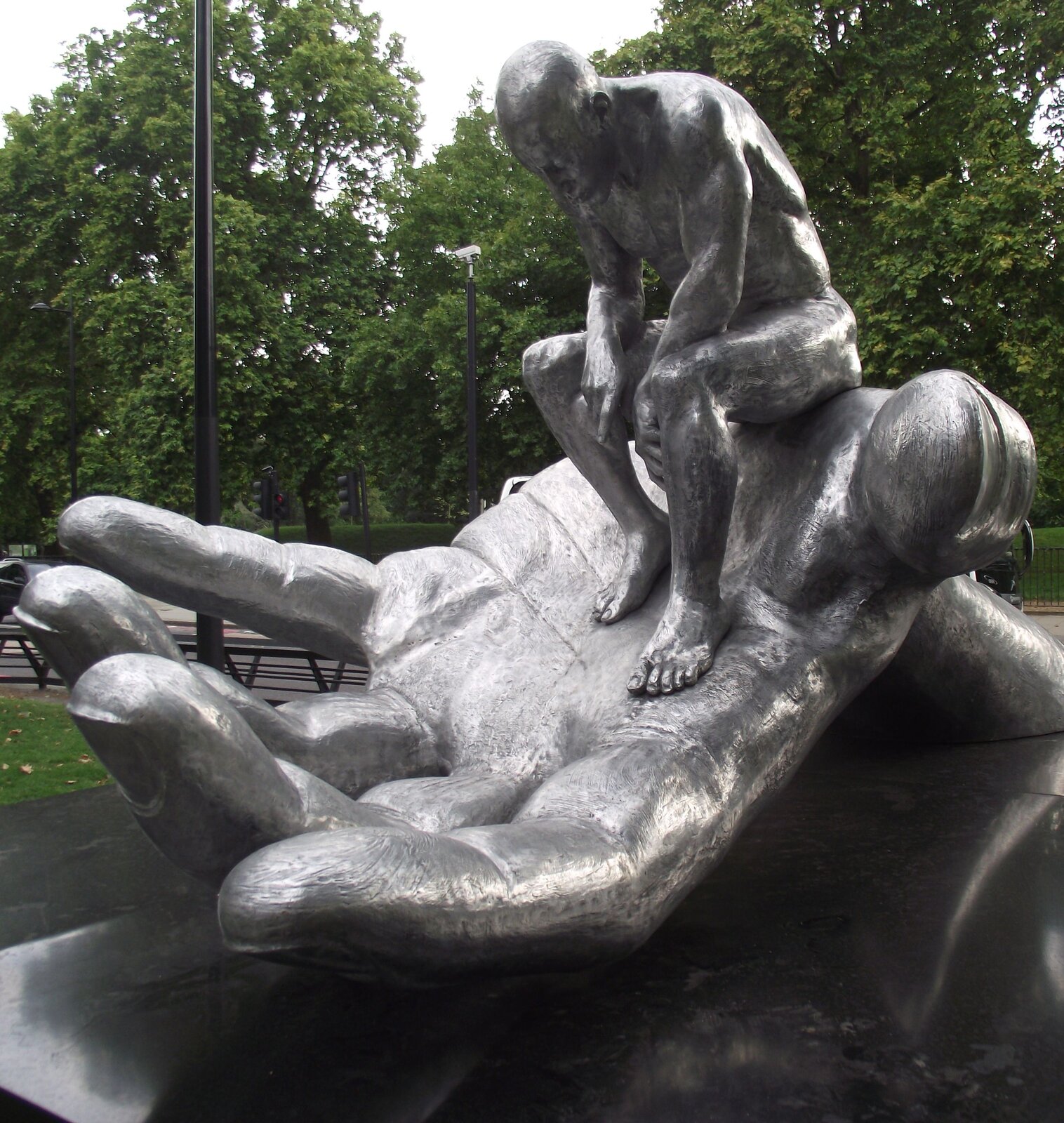 Zdjęcie wykonane w pochmurny dzień przedstawia instalację  Lorenza Quinna pod tytułem „Ręka Boga”. Praca prezentuje postać nagiego mężczyzny siedzącego na gigantycznej dłoni. Mężczyzna jest łysy, pochylony do przodu, jego głowa skierowana jest do dołu. Oczy ma zamknięte, dłonie opiera o łydki. Postać jak i dłoń są w srebrnym kolorze. Instalacja znajduje się w parku miejskim, o czym świadczą stojące ławki przy ścieżkach oraz duża ilość drzew.