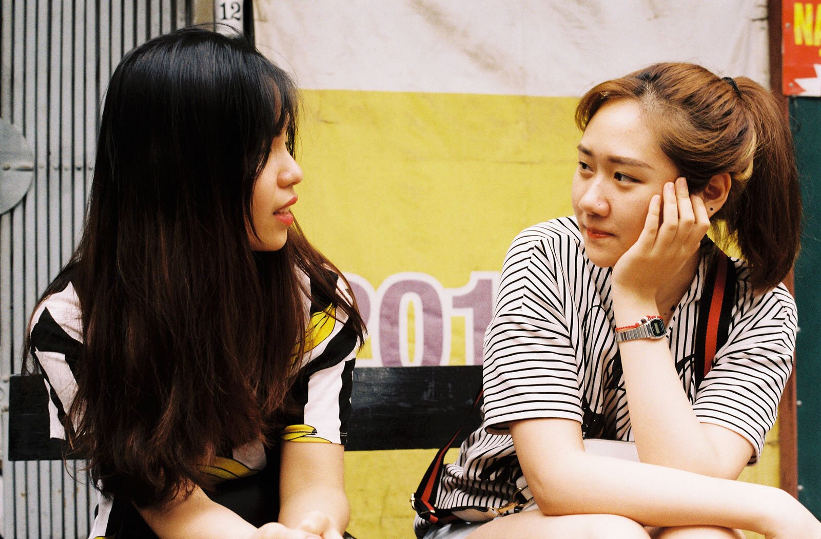 Zdjęcie przedstawia dwie nastolatki siedzące na ławce i patrzące na siebie. Dziewczyna po lewej stronie ma długie, ciemne włosy opadające na ramiona, jest ubrana w kolorową bluzkę z krótkim rękawem, a jej ręce są wyciągnięte do przodu i złożone na kolanach. Głowę ma zwróconą w prawą stronę. Dziewczyna po prawej ma długie, rude włosy związane w kucyk, jest ubrana w bluzkę z krótkim rękawem w biało‑czarne paski. Zwróconą w lewo głowę podpiera lewą ręką, a prawą rękę ma położoną na kolanach. W tle widoczne są kolorowe transparenty.