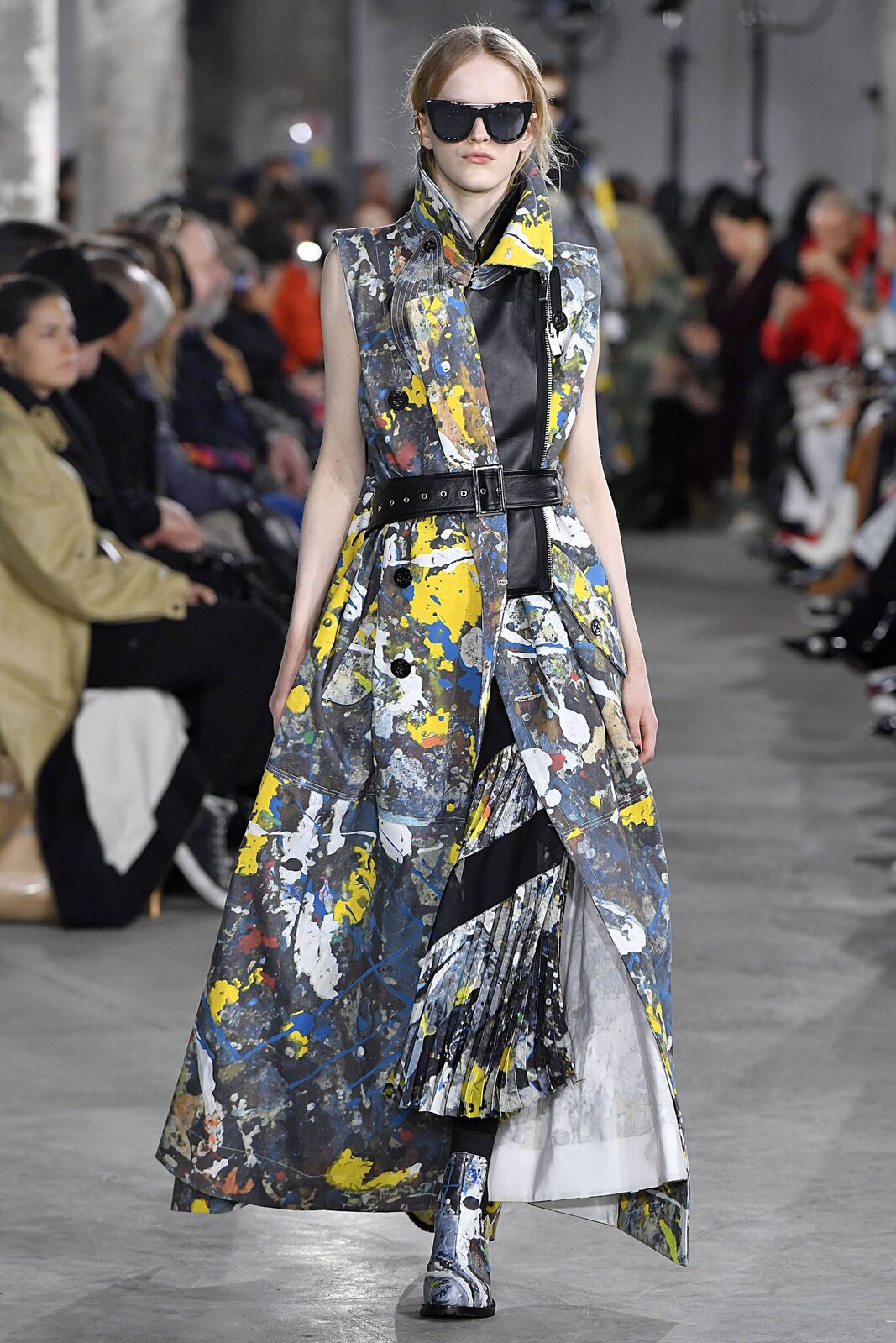 Fotografia przedstawia fragment pokazu mody – na wybiegu pomiędzy widownią przechadza się młoda szczupła blond włosa kobieta ubrana w czarny płaszcz, sukienkę oraz buty, które sprawiają wrażenie pobrudzonych farbą – są to elementy garderoby, w których projektant mody zainspirował się malarstwem abstrakcyjnym.