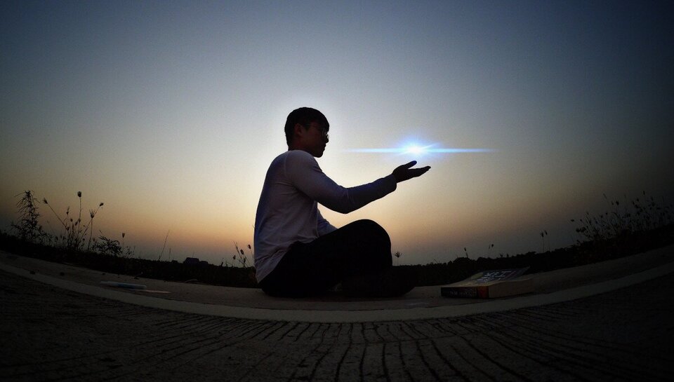 Sylwetka chłopca siedzącego po turecku na betonowej powierzchni o świcie. Postać trzyma wyciągniętą dłoń, nad którą widoczny jest jasny punkt światła przypominający małą gwiazdę lub refleks. W tle rozjaśniające się niebo oraz zarys roślinności i linii horyzontu. Na ziemi obok chłopca leży książka.