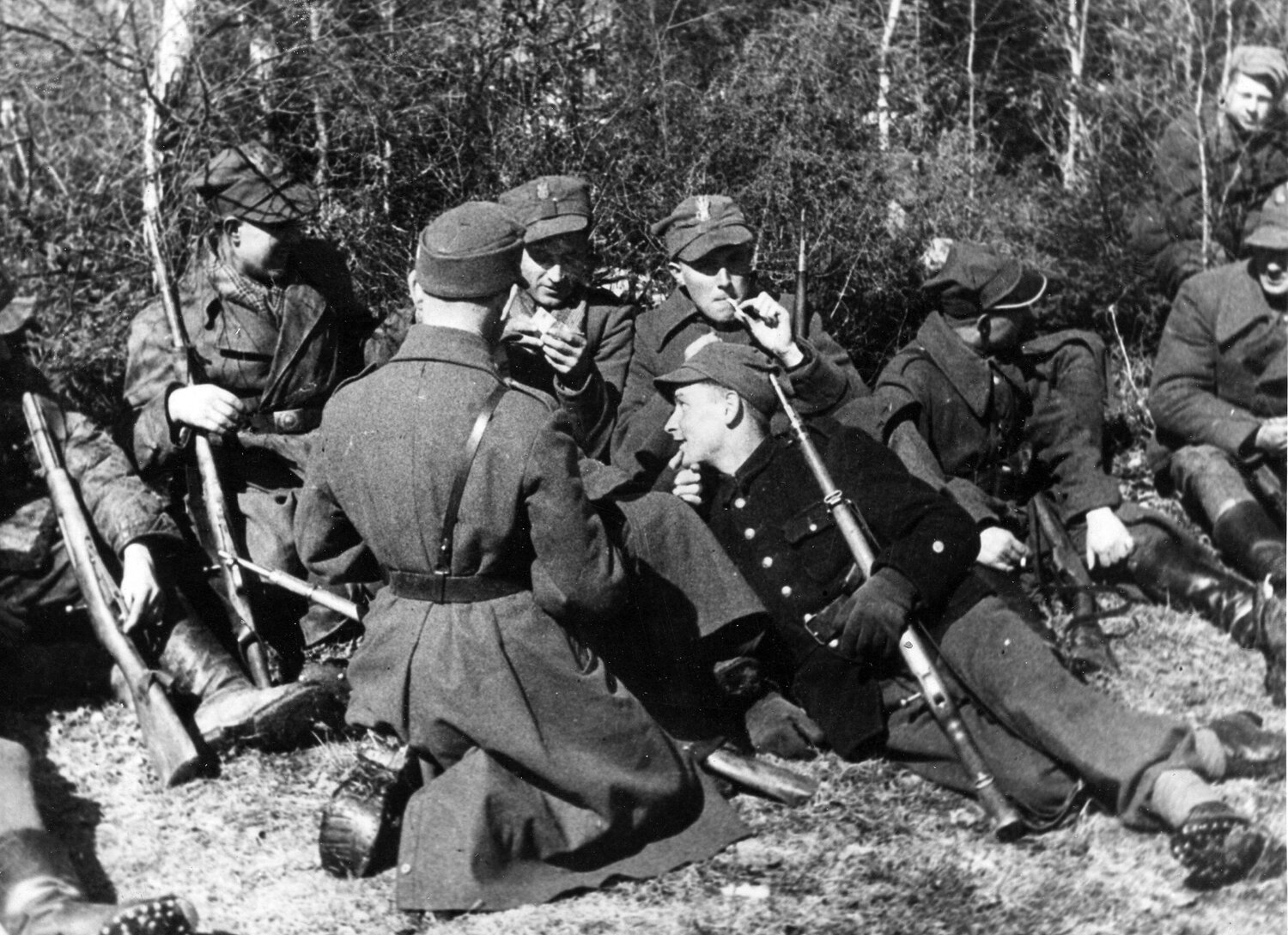 Zdjęcie przedstawia leżących na trawie żołnierzy. Trzymają broń i rozmawiają ze sobą, kilku pali papierosy. Są ubrani w proste, długie mundury oraz czapki z orłem. Za nimi jest las.