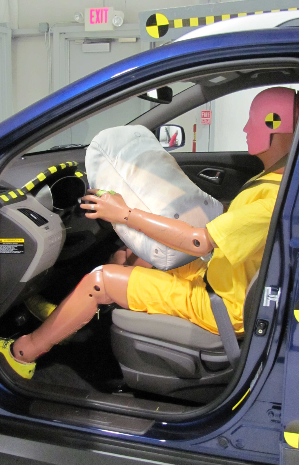 Zdjęcie przedstawia próbę uderzeniową, jakiej poddaje się samochody. Ukazano widok z perspektywy osoby stojącej przy drzwiach kierowcy. Ubrany na żółto manekin testowy, przypięty do fotela kierowcy ma przed sobą wystrzeloną białą poduszkę powietrzną.