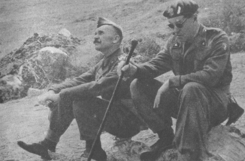 Fotografia przedstawia dwóch mężczyzn ubranych w wojskowe mundury. Siedzą na kamieniach, jeden patrzy w niebo, drugi w ciemnych okularach patrzy w ziemię i trzyma laskę w ręku. W tel widać skalisty teren porośnięty trawą.