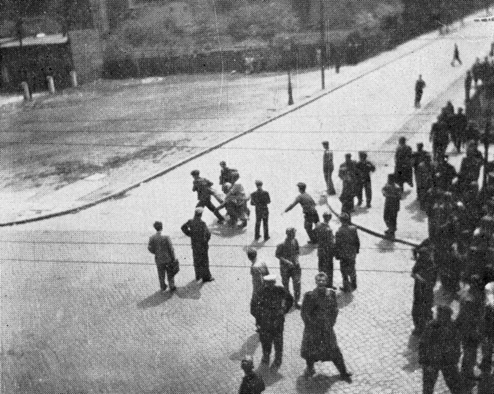 Zdjęcie przedstawia tłum ludzi na ulicy podczas starcia z policją. Grupa wynosi rannego mężczyznę.