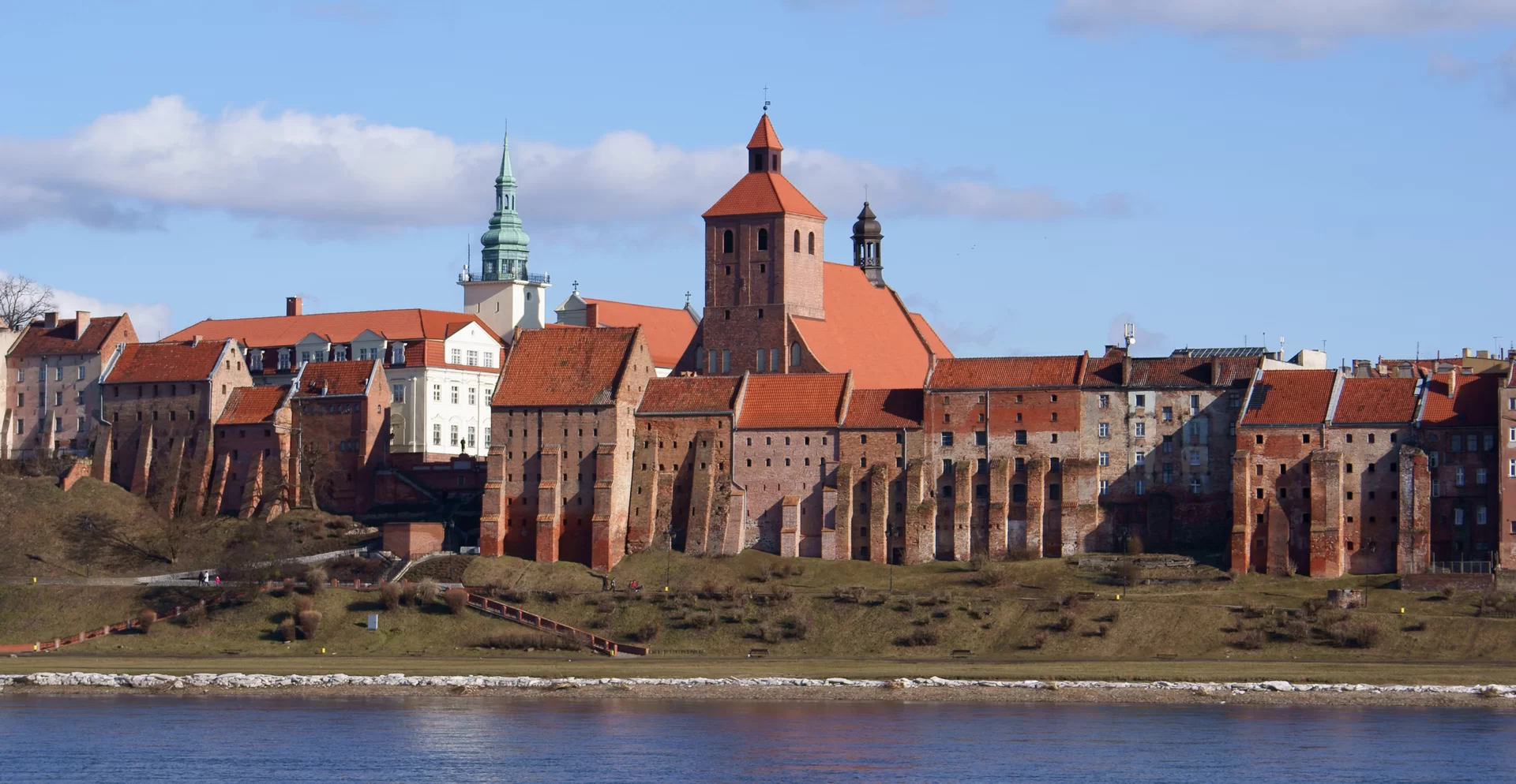 Kliknij, aby powiększyć Zdjęcie przedstawia widok na miasto nad rzeką. Spichlerze znajdują się na pierwszym planie, stoją blisko brzegu rzeki. Są to wysokie budynki z małymi oknami. Na drugim planie widać dachy zabudowań.
