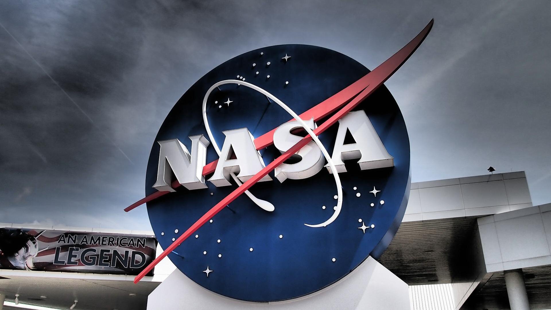 Grafika przedstawia logo NASA. Logo NASA to okrąg, wewnątrz którego umieszczono napis „NASA”, gwiazdy, linię oraz ikonę w kształcie litery „V”.