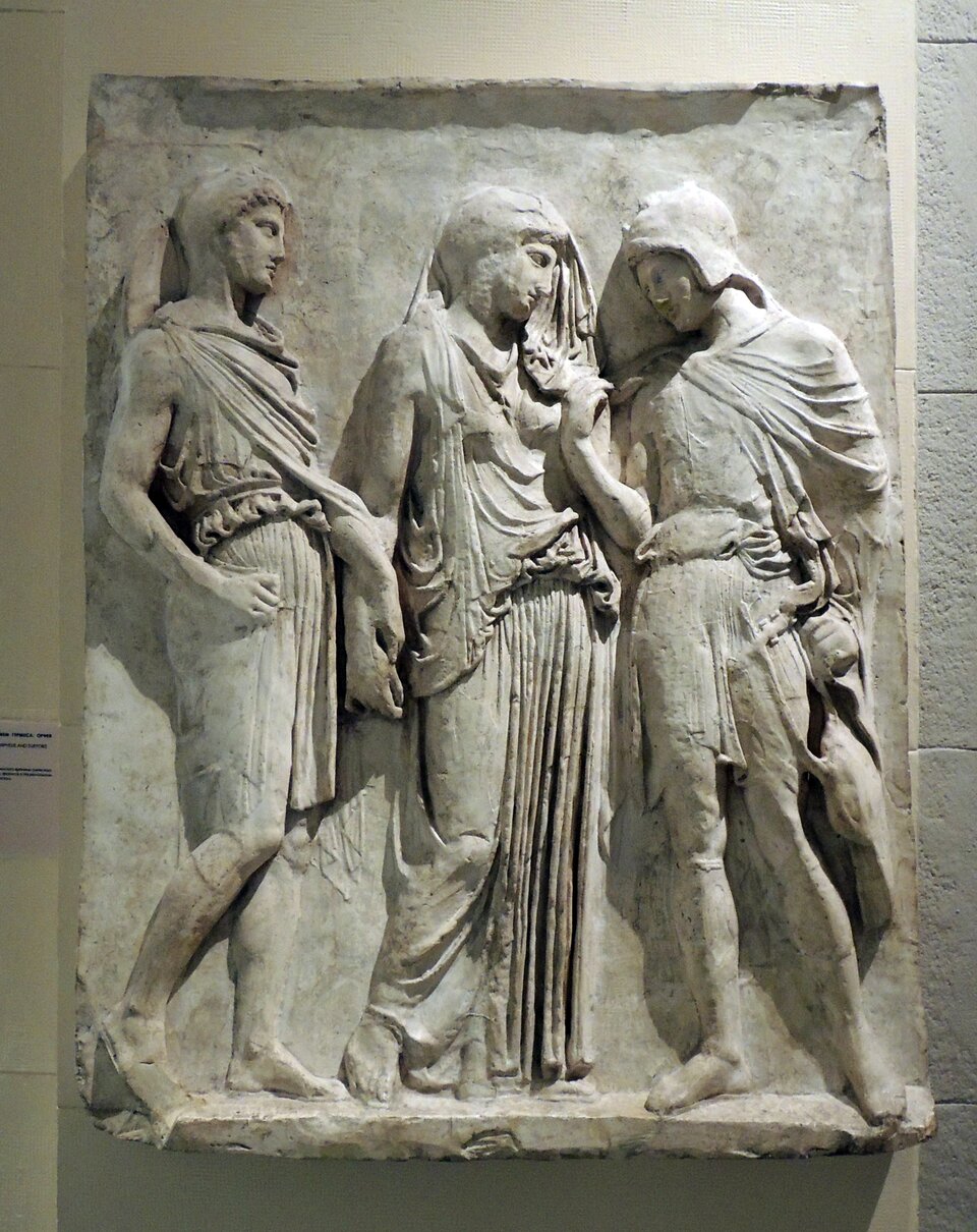 Zdjęcie przedstawia starożytną płaskorzeźbę. Przedstawia ona dwóch mężczyzn i stojącą pomiędzy nimi kobietę.