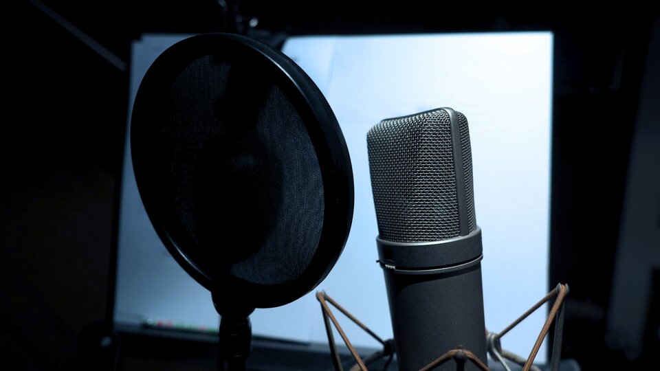 Mikrofon pojemnościowy oraz pop filter Z prawej strony fotografii znajduje się duży, szary mikrofon z brązowymi elementami na dole. Z lewej strony jest tytułowy pop filtr. Jest on duży, okrągły i w czarnym kolorze. W tle obrazka jest rozmyty, jasny ekran.