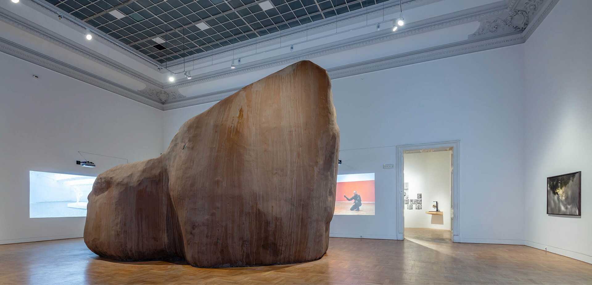 Na zdjęciu znajduje się pomieszczenie o brązowej podłodze, białych ścianach i czarnym suficie. Z sufitu zwisa wiele małych lampek. Na środku fotografii znajduje się wielka rzeźba o nieregularnym kształcie i kolorze beżowo‑brązowym. Za rzeźbą na ścianach wiszą 3 obrazy. Są one nieostre, niewyraźne. Pomiędzy drugim a trzecim obrazem jest przejście do kolejnego pomieszczenia. W tle jest kolejne pomieszczenie z obrazami na białych ścianach. 