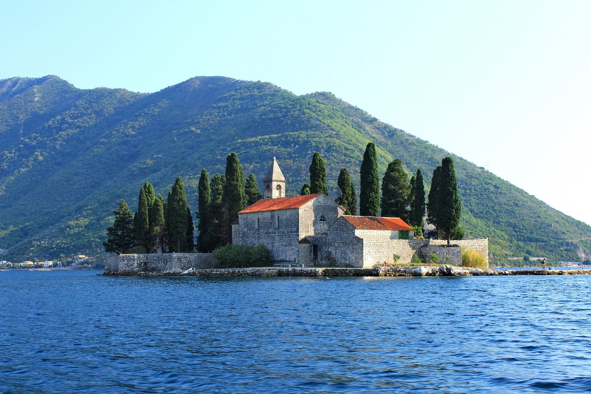 Ilustracja przedstawia wyspę, na której wznosi się niewielki kościółek i rosną cyprysy.