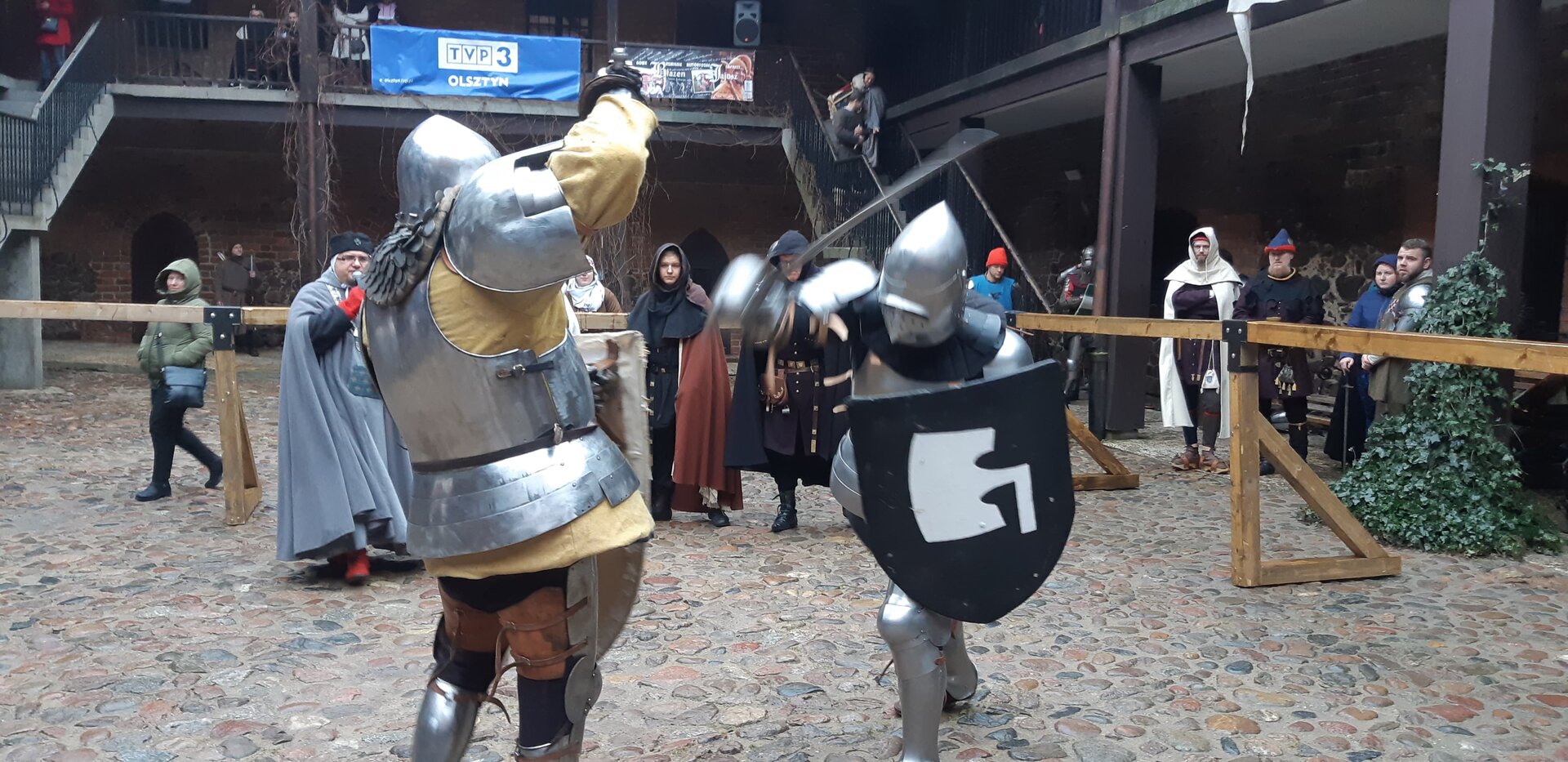 Fotografia przedstawia turniej rycerski na Zamku w Nidzicy. Na środku zdjęcia znajdują się osoby ubrane w zbroje rycerskie, które walczą ze sobą. Za walczącymi rycerzami po prawej stronie znajdują się ludzie ubrani w rycerskie stroje patrzący na widowisko. W tle znajdują się schody prowadzące na balkon, z którego ludzie podziwiają spektakl.