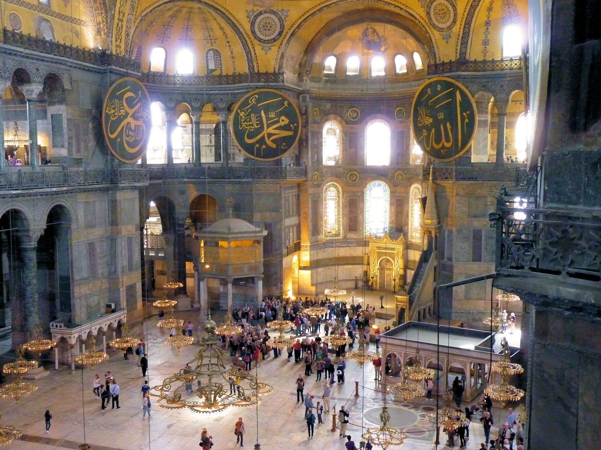 Zdjęcie współczesne, przedstawiające wnętrze meczetu Hagia Sophia. Zdjęcie wykonane z wysokości piętra przedstawia widok na nawę główną świątyni. W części parterowej widać pojedyncze osoby spacerujące po świątyni, a także grupy turystów zwiedzających obiekt. W lewej części fotografii widoczne przejście do nawy bocznej - fragment arkady, widoczne biforium na piętrze wyżej i arkady przez które pada światło dnia. Widoczne medaliony z wezwaniami modlitwy w języku arabskim. Widoczny fragment sklepienia dekorowany motywami orientalnymi. Wnętrze konstrukcji wykonane z alabastru i granitu, tonacja kolorów sklepienia utrzymana w ciepłym odcieniu żółtego. 