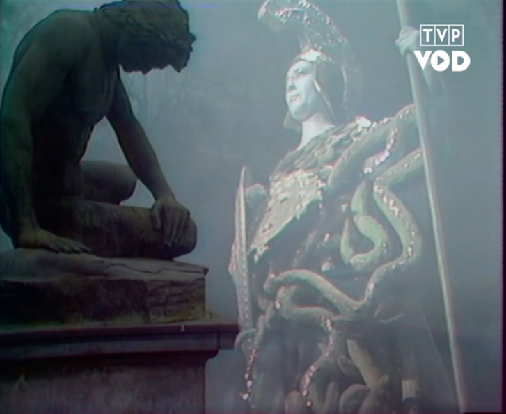 Fotografia przedstawia nakładające się na siebie dwa obrazy. Po lewej stronie ukazana jest rzeźbiona postać młodego nagiego mężczyzny. Postać ukazuje prawy profil. Mężczyzna ma gęste włosy, gładkie czoło, prosty nos i niewielkie usta. Pochyla głowę. Siedzi z nogami ugiętymi w kolanach, opierając się o prawą rękę. Po prawej stronie stoi kobieta w sile wieku. Postać jest ukazana z perspektywy od dołu. Kobieta prezentuje lewy profil. Ma gładkie czoło, ciemne brwi, lekko zwężone oczy, niewielki nos i wydatne usta. Patrzy przed siebie. Jest ubrana w zbroję. Na głowie ma hełm. W prawej dłoni trzyma tarczę, a w lewej długą włócznię.