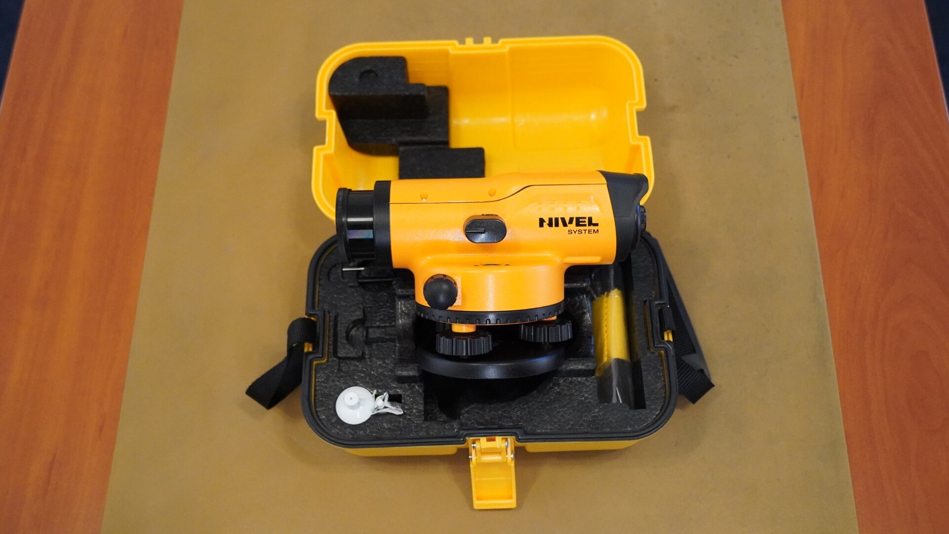 Na zdjęciu zaprezentowano niwelator umieszczony w walizce służącej do jego przenoszenia. Instrument posiada czarną okrągłą podstawę – spodarkę, na niej znajdują się śruby, a na nich korpus z lunetą, która z jednej strony ma okular, a z drugiej obiektyw. Widoczne jest również jedno pokrętło. Korpus urządzenia jest w kolorze żółtym, a podstawa, śruby, pokrętła oraz elementy obudowy są czarne. 