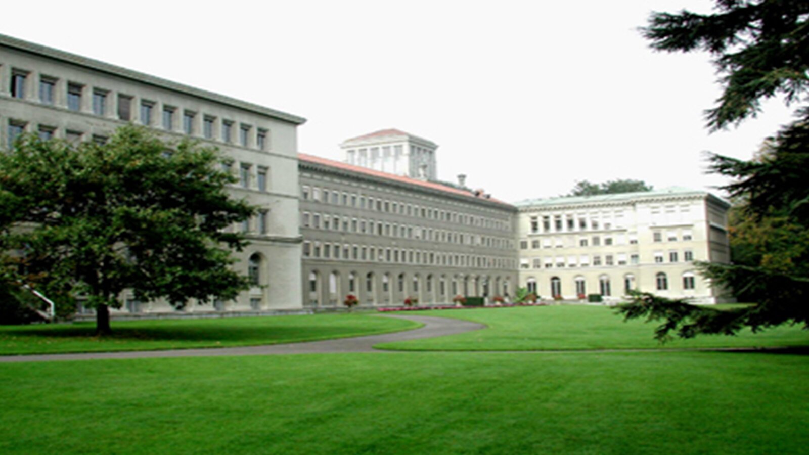 Zdjęcie przedstawia kompleks budynków zbudowanych z jasnego kamienia, z dużą ilością okien i drzwi wejściowych. Dolne okna mają półokrągłe framugi. Przed nimi i po prawej stronie widoczną są fragmenty zielonych drzew oraz wypielęgnowany trawnik.