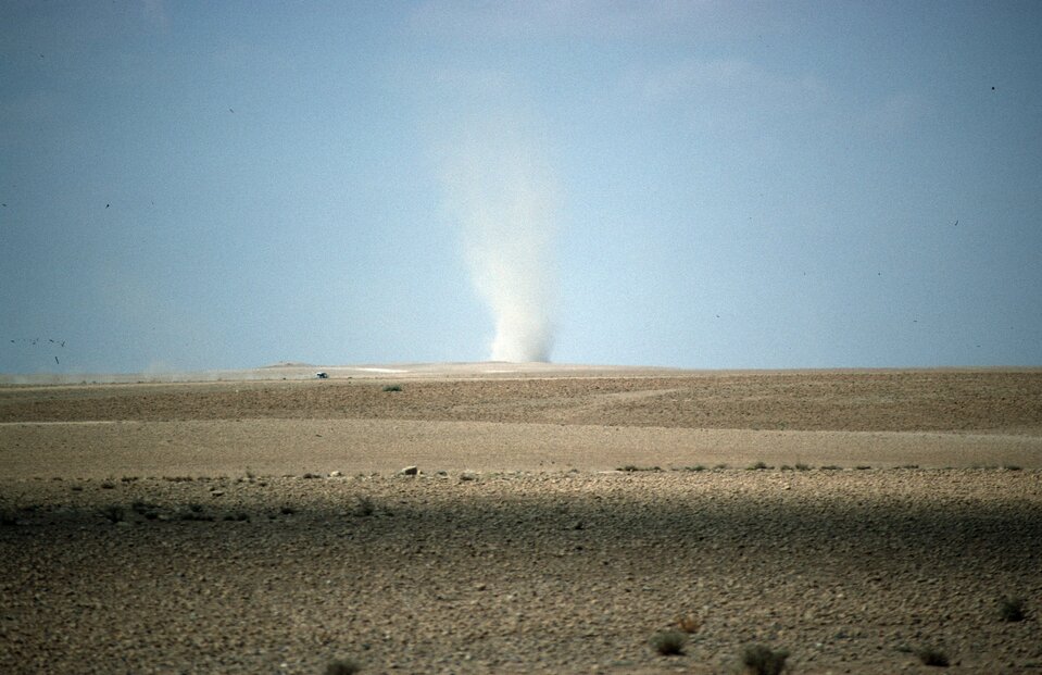 Na zdjęciu jest dość płaski teren, pustynny. Na linii horyzontu znajduje się biały, szeroki wir powietrza, unoszący się ku górze. Przypomina szeroki słup dymu.