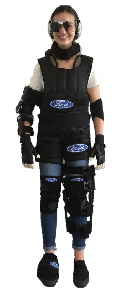 Na zdjęciu przedstawiono kobietę ubraną w symulator wieku. Założone ma specjalistyczne słuchawki oraz okulary, a na ciele ma ubrane liczne taśmy, stelaże i kamizelkę obciążającą.