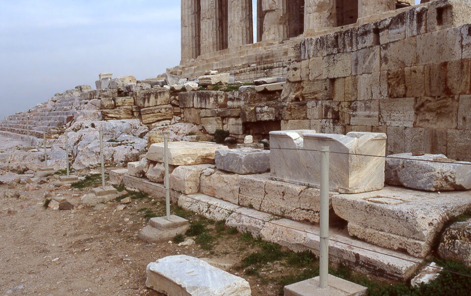 Fragmenty Starej Świątyni Ateny na Akropolu. Liczne rozrzucone w nieładzie bloki skalne, na dalszym planie widoczne fragmenty kolumn.