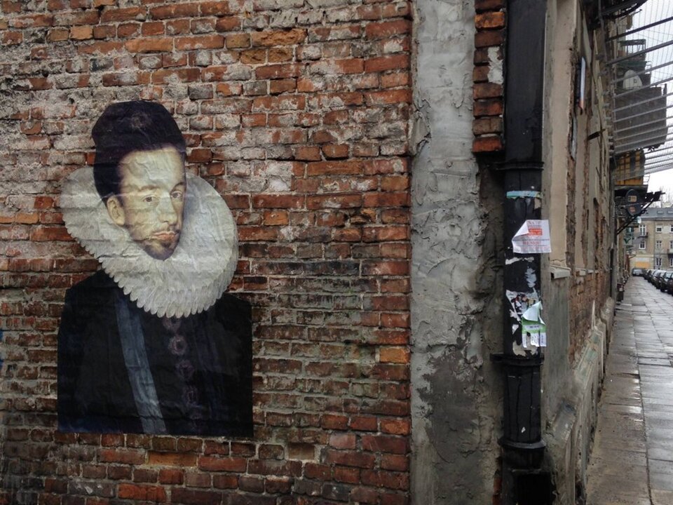 Ilustracja przedstawia mural wykonany na murze z czerwonej cegły. Widoczny jest mężczyzna przedstawiony od piersi w górę. Ubrany jest w czarny strój z dużymi guzikami, wokół szyi ma białą kryzę. Czarne włosy ma upięte wysoko. Jego twarz jest pociągła z lekko zarysowanym ciemnym zarostem.