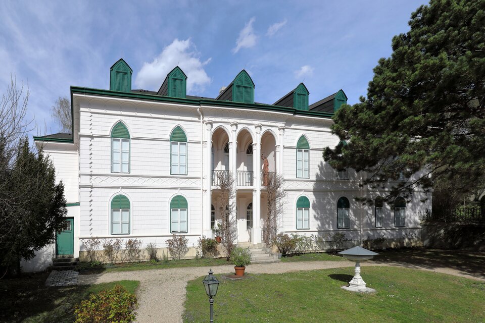 Zdjęcie przedstawia willę. Przed nią znajduje się ścieżka usypana ze żwiru oraz trawnik z fontanną dla ptaków. Budynek jest biały. Jego okiennice, drzwi i dach są zielone. Nad wejściem są kolumny. Z prawej strony rośnie rozłożyste drzewo.