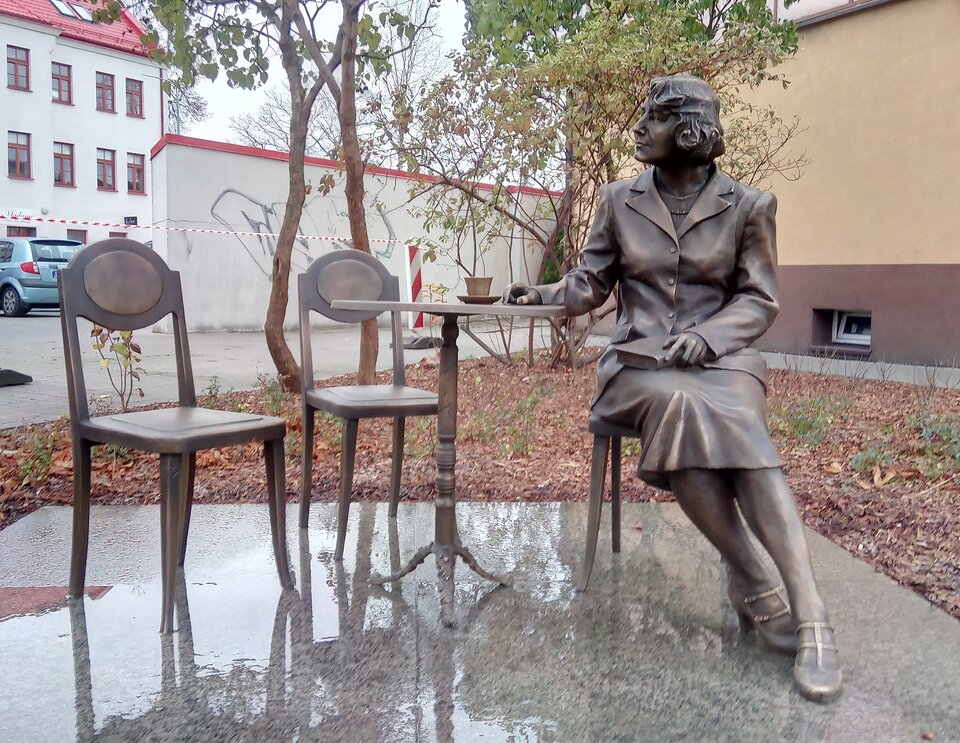 Fotografia przedstawia pomnik. Stanowią go trzy krzesła i stolik kawowy, na którym stoi filiżanka postawiona na spodku. Dwa krzesła stojące po lewej stronie są puste. Na krześle po prawej stronie siedzi kobieta w sile wieku. Postać ukazuje lewy profil. Ma owalną twarz, prosty nos i wąskie usta. Jej włosy lekko falują, sięgają żuchwy. Kobieta jest ubrana w koszulkę i zapięty żakiet oraz spódnicę sięgającą za kolano. Na stopach ma pantofle. Prawą rękę opiera o stolik. W lewej trzyma książkę opartą o prawe udo. W tle stoją miejskie zabudowania.