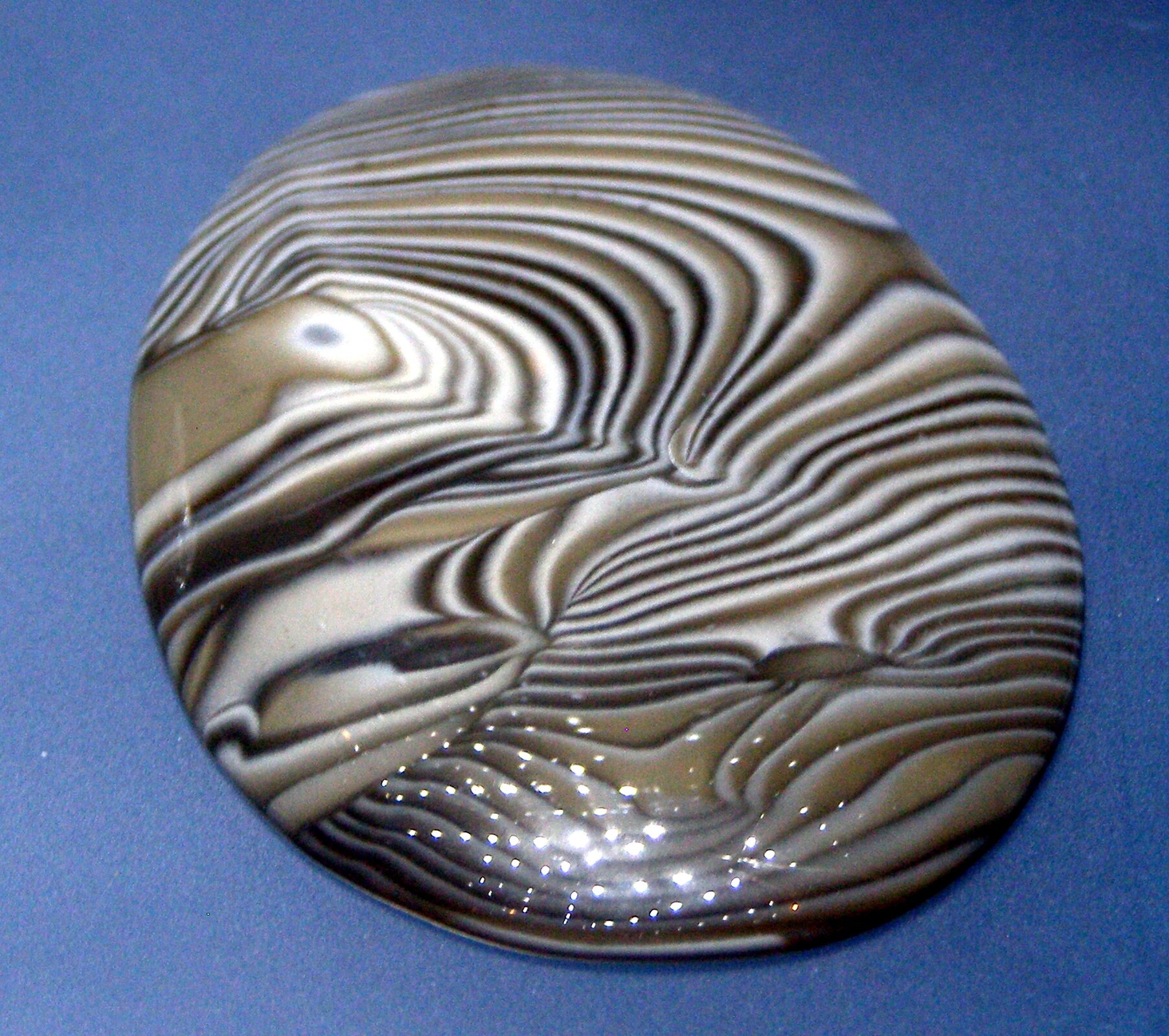 Zdjęcie przedstawia okrągły, gładko oszlifowany kawałek krzemienia pasiastego na niebieskim tle. Kamień cały pokryty jest nieregularnym wzorem składającym się z powyginanych pasków, naprzemiennie czarnych, białych i brązowych. Kamień błyszczy, świadczą o tym widoczne w dolnej jego części odblaski lampek oświetlających ekspozycję.