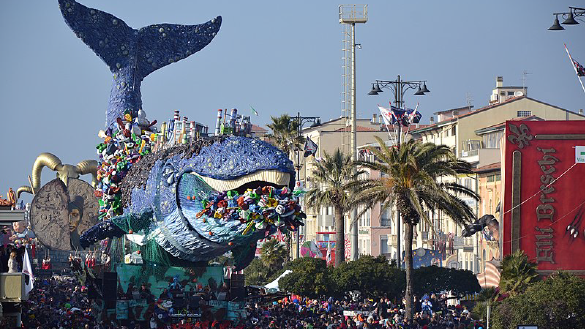 Zdjęcie przedstawia paradę w trakcie karnawału w Viareggio. Zdjęcie wykonane jest w słoneczny dzień. Wzdłuż ulicy, po której jadą   karnawołowe platformy rosną zielone drzewa i wysokie palmy. Na ulicy przemieszcza się tłum kolorowo ubranych ludzi. Dużą część kadru zajmuje platforma z wielorybem, któremu z pyska "wypadają" barwne figury przypominające ludzi. Takie same znajdują się również na wielorybie.  