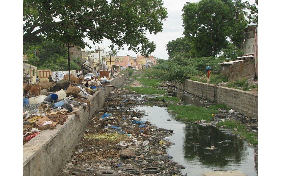 Zdjęcie przedstawia zanieczyszczenie wody w Indiach. Na zdjęciu kanał wodny biegnący wzdłuż osiedla, w którym pełno jest śmieci, opon samochodowych, foliówek, plastikowych rur, i tak dalej. 