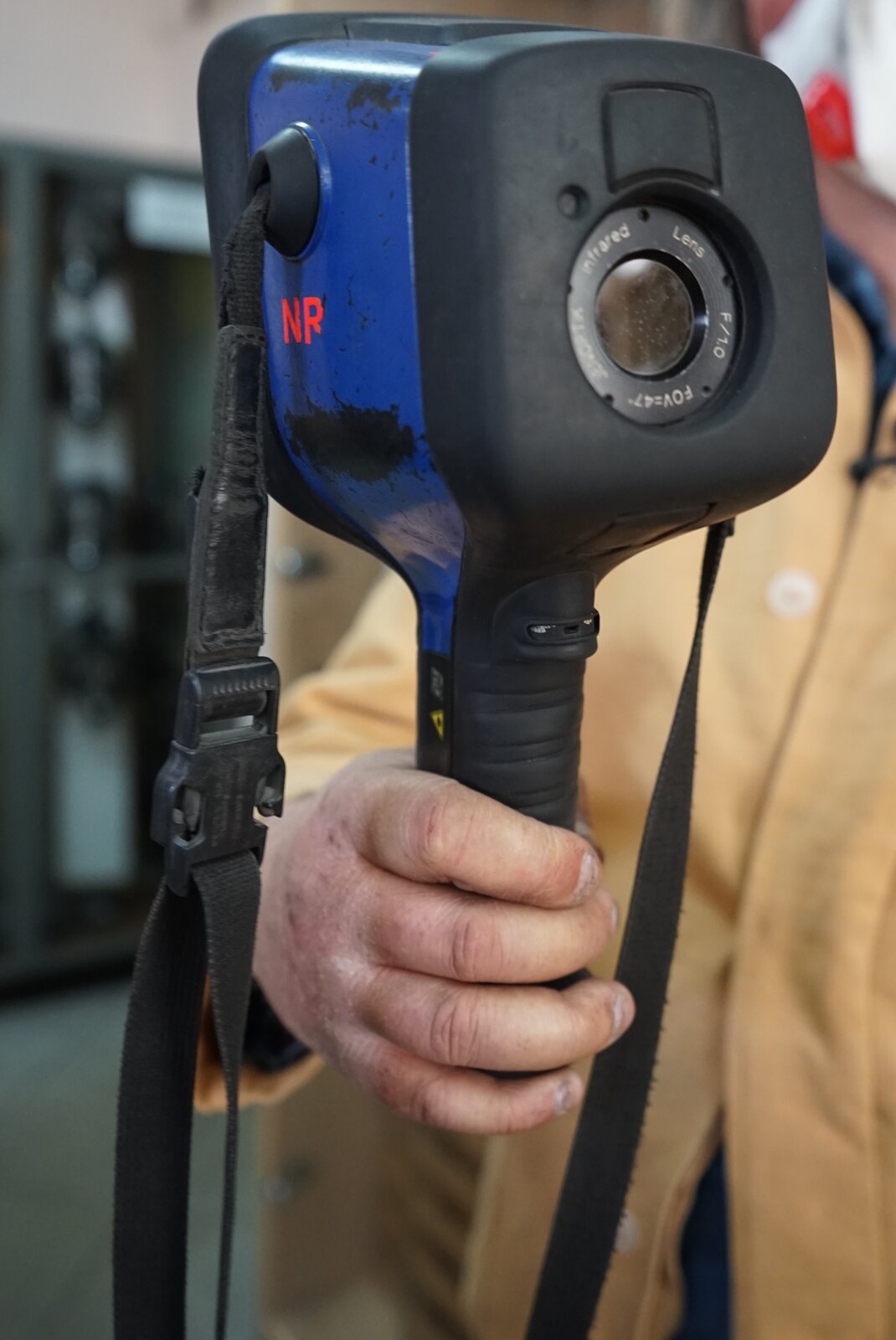 Zdjęcie przedstawia kamerę termowizyjną. Kamera osadzona jest na uchwycie pistoletowym. W górnej, prostopadłościennej części wbudowany jest okrągły obiektyw. Do obudowy kamery przypięty jest pasek. Kamera jest prezentowana w ręce przez górnika. Górnik ubrany jest w strój ochronny.