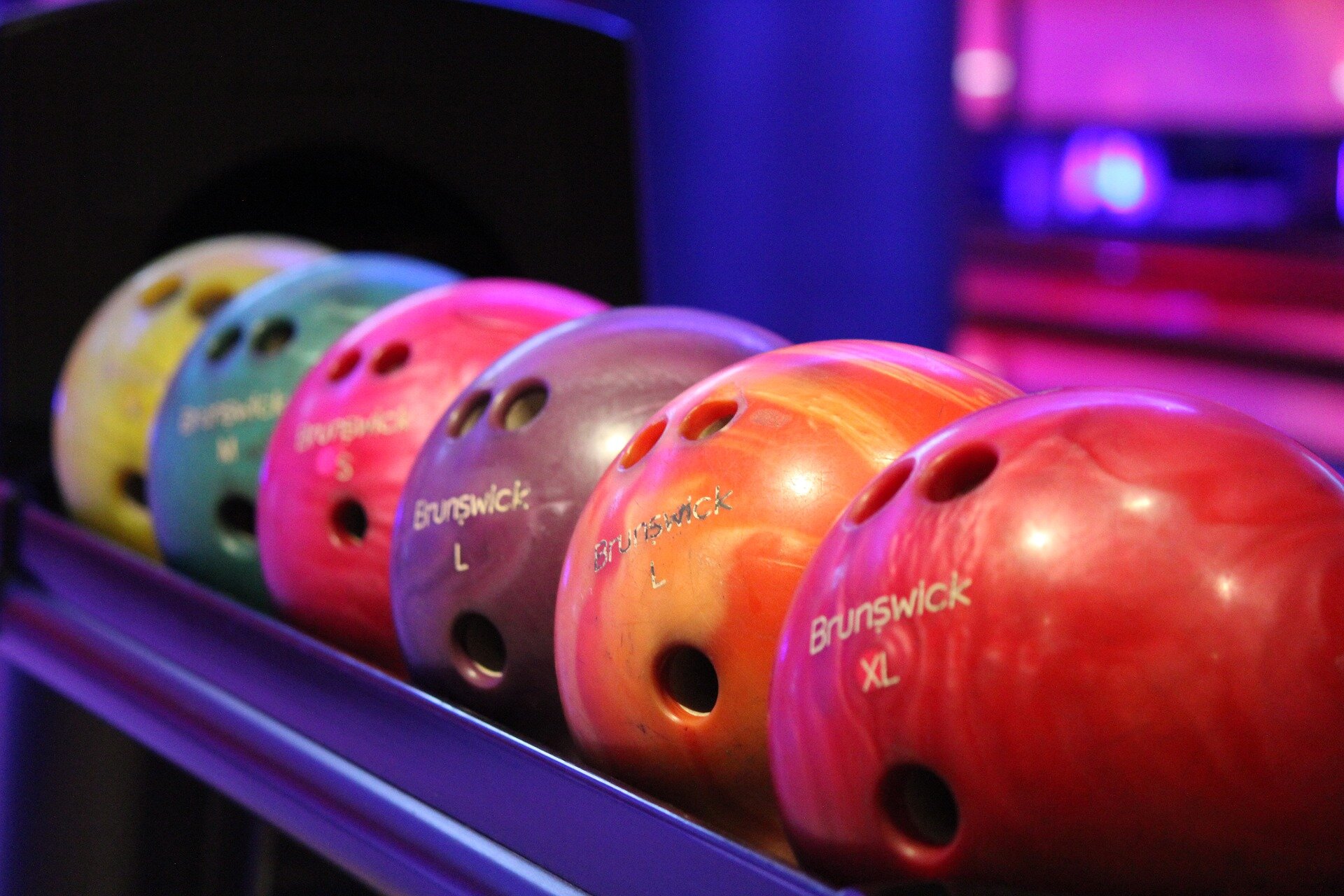 Rys. a. Zdjęcie przedstawia kolorowe kule (bowling) do zbijania kręgli, ułożone na półce. W kulach widoczne są otwory/uchwyty na palce. 