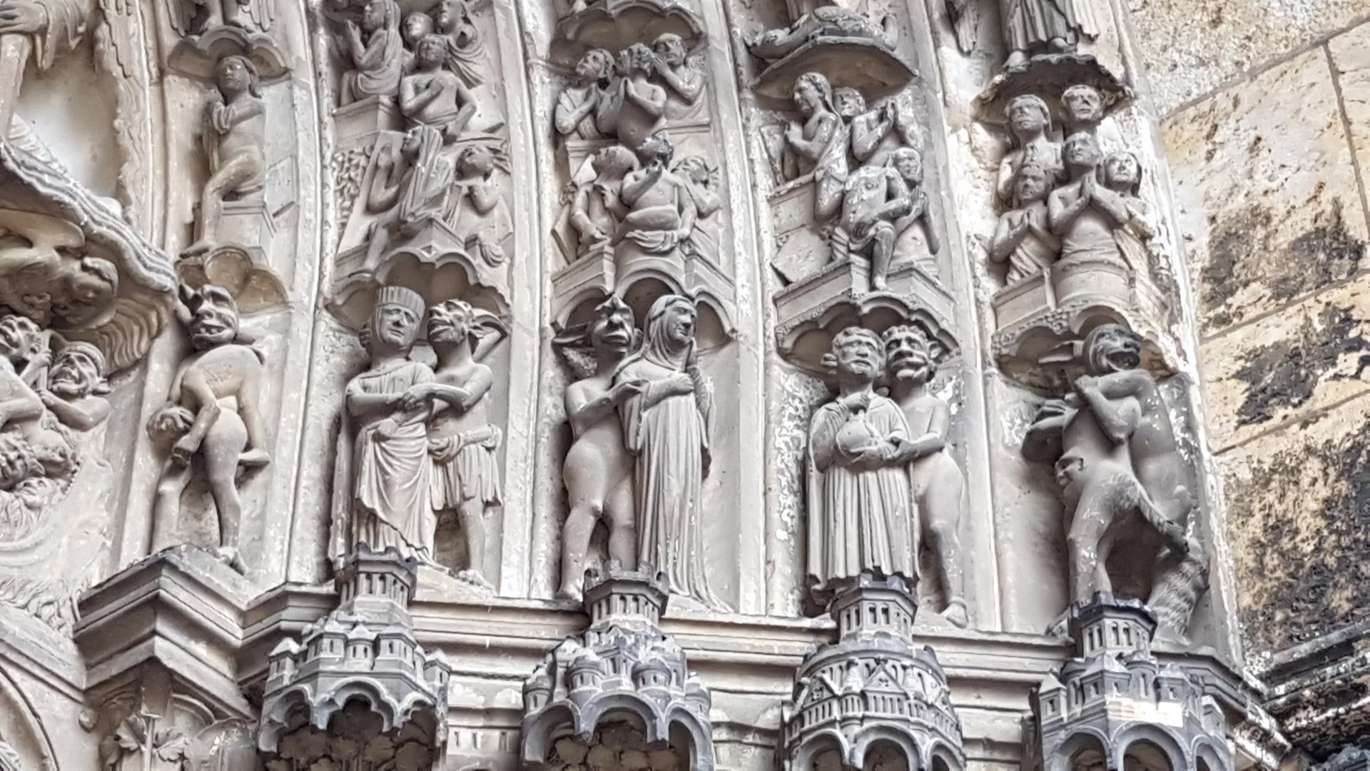 Zdjęcie przedstawia fragment portalu. Portal składa się z długich pasów, z których każdy kolejny wychodzi dalej z portalu. Pasy tworzą schodkowe uskoki. Każdy taki pas składa się z rzeźb, ustawionych jedna na drugiej. Są to w większości stojące postacie. Jedne przypominają ludzi, a drugie to stworzenia z długimi uszami i dużymi zębami.