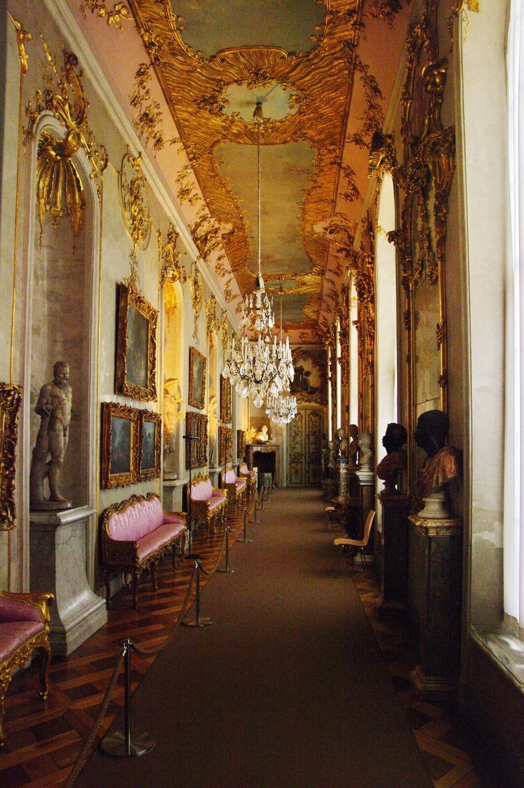 Ilustracja przedstawia wnętrze pałacyku Sanssouci w Poczdamie. Ilustracja ukazuje korytarz. Po jednej stronie znajdują się okna, między zbiorami są popiersia na kolumnach. Po prawej stronie, na ścianie wiszą obrazy, między zbiorami są sofy i kolumny. Na kolumnach stoją rzeźby. Sufit pałacu jest bogato zdobiony w ornamenty oraz malowidła.