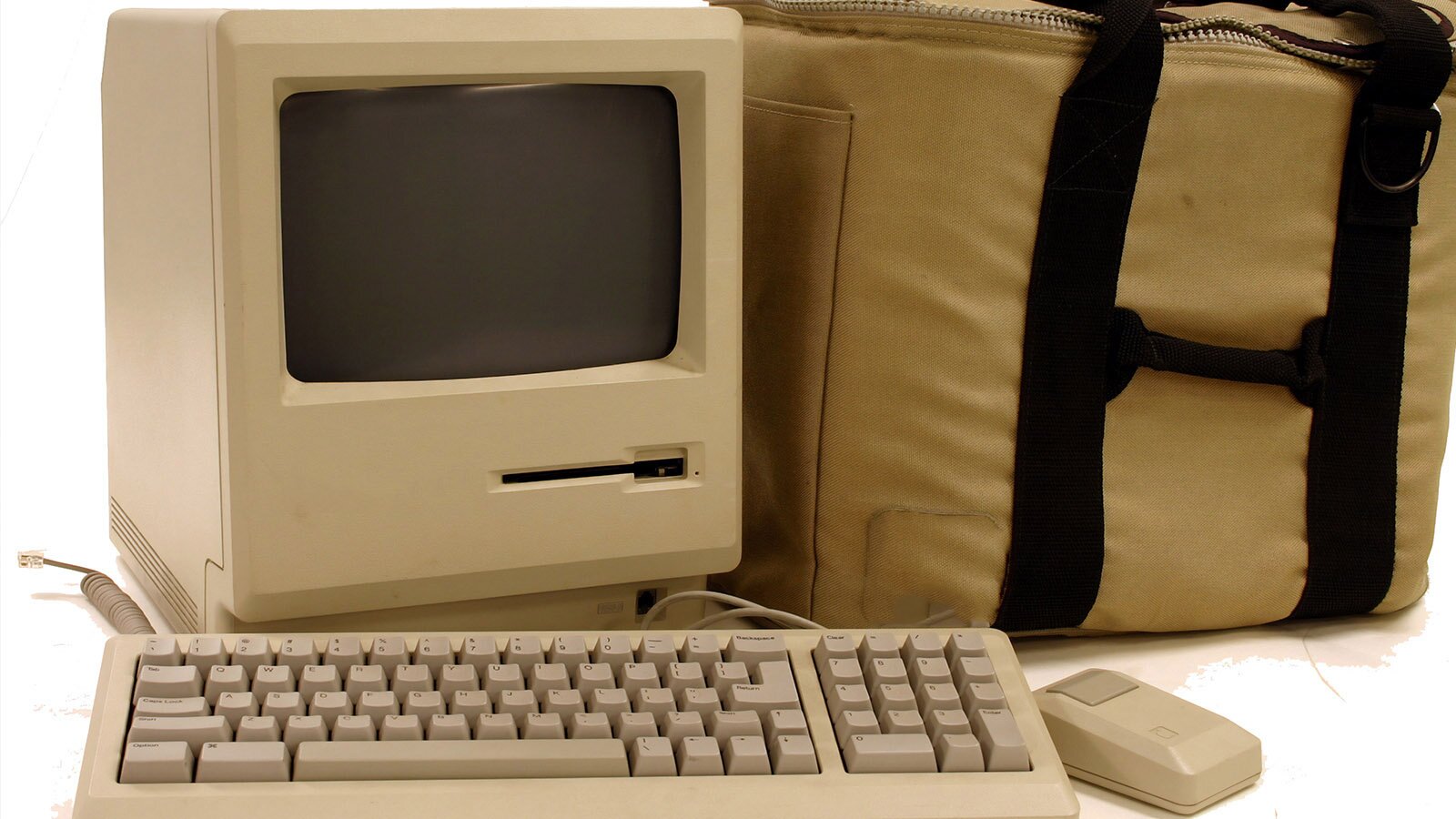 Zdjęcie przedstawia jeden ze starych modeli komputera stacjonarnego. Widoczny jest masywny monitor z małym ekranem, klawiatura, prostopadłościenna myszka oraz duży kremowy pokrowiec na komputer.