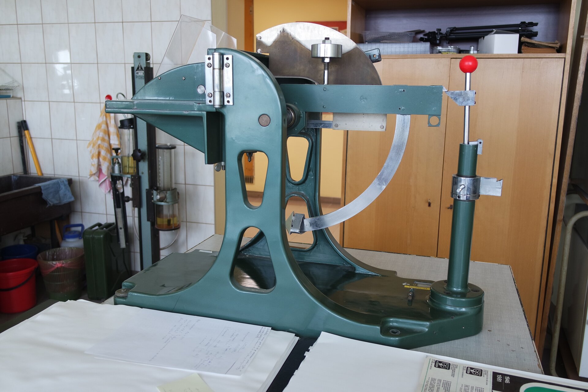 Zdjęcie prezentuje aparat do oznaczania odporności tektury falistej na przebicie z głowicą w kształcie ostrosłupa. Urządzenie zbudowane jest z głowicy w kształcie ostrosłupa, mechanizmu wyzwalającego (zakończonego czerwoną kulką) i skali do odczytania wyniku.