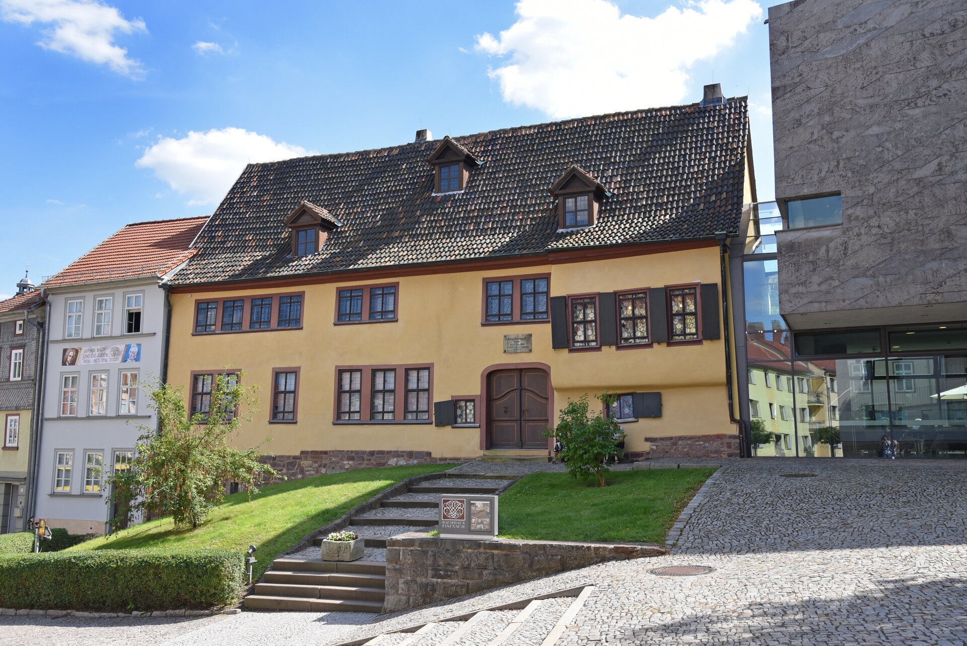 Fotografia przedstawia dom w Eisenach, w którym mieszkał młody Jan Sebastian Bach. Na fotografii widać żółty duży dom. Główne miejsce zajmuje wejście do budynku. Okna mają brązowe obramowania i okiennice. Przed domem widać kamienną ławkę, schody i zieleń.