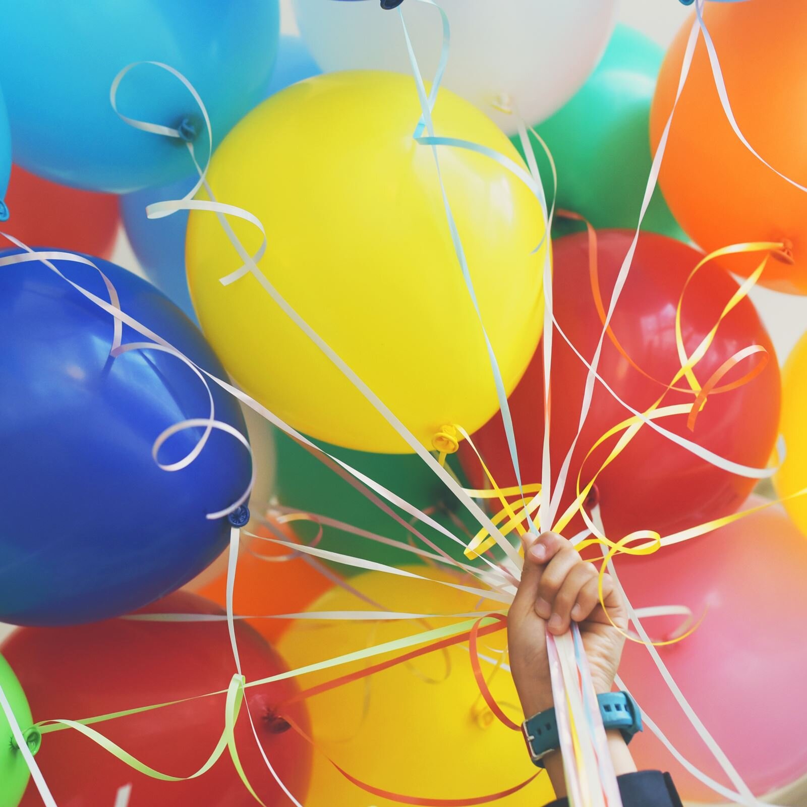 Zdjęcie przedstawia lewą dłoń trzymającą pęk kolorowych balonów przywiązanych tasiemkami, pomiędzy którymi widoczne są kolorowe serpentyny.