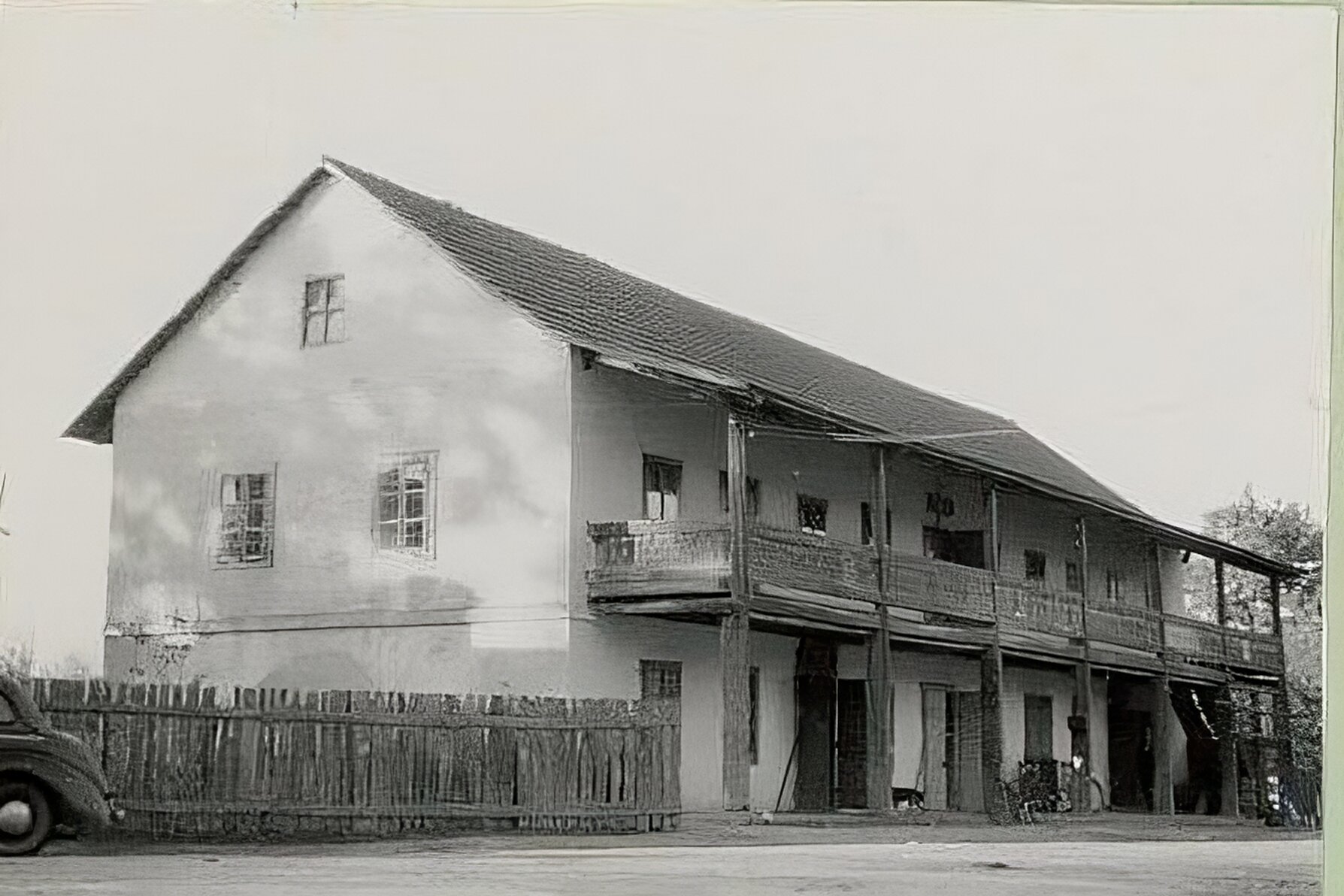 Zdjęcie przedstawia dwukondygnacyjny murowany budynek papierni z drewnianym balkonem biegnącym wzdłuż  piętra budynku. Balkon wsparty jest w siedmiu miejscach grubymi, drewnianymi podporami. Drewniana  balustrada jest rzeźbiona. Budynek jest przykryty dwuspadowym dachem. Z lewej strony widać płot i tył stojącego przed nim samochodu. Obok budynku z prawej strony rosną drzewa.