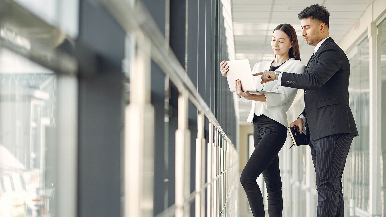 Zdjęcie przedstawia kobietę i mężczyznę stojących obok siebie na korytarzu, przy ścianie wypełnionej oknami. Oboje są ubrani na biało czarno. Mężczyzna ma na sobie garnitur, w prawej ręce trzyma notes, a lewą ręką wskazuje na ekran laptopa, trzymanego przez kobietę. Kobieta ma na sobie czarne spodnie oraz biały żakiet. 