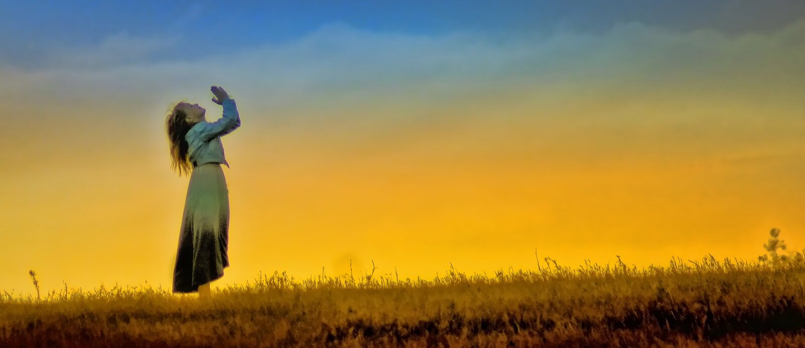 Zdjęcie przedstawia kobietę stojącą na łące z rękoma wzniesionymi do nieba w modlitwie. Niebo ma niebiesko‑żółtą barwę.