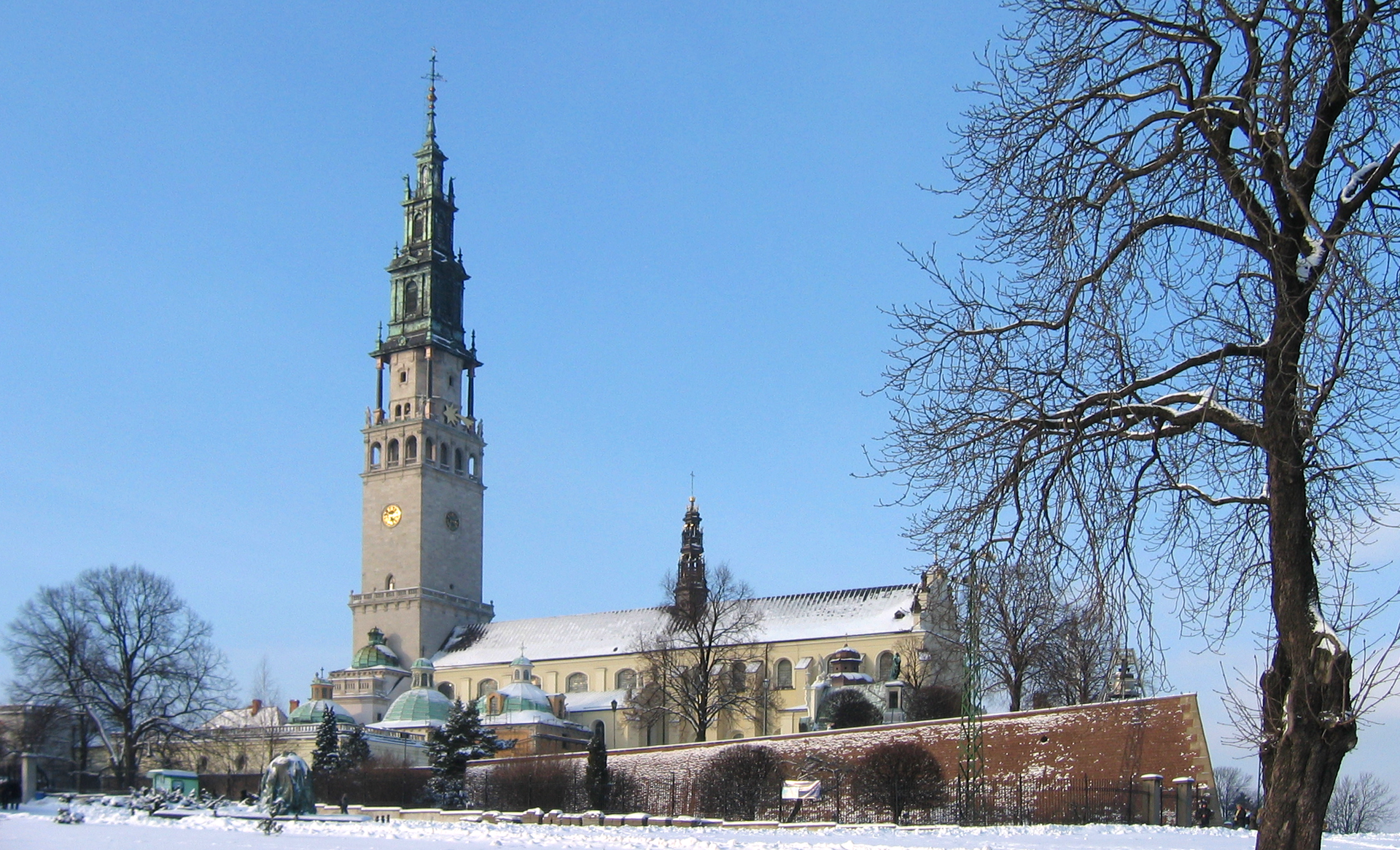 Fotografia barwna przedstawiająca klasztor zakonu paulinów w Częstochowie w zimowej szacie. Po lewej stronie budowli jest strzelista wieża, na jej czubku widać krzyż. Color photography depicting the monastery of the Pauline order in Częstochowa in a winter robe. On the left side of the building there is a towering tower with a cross on its top.
