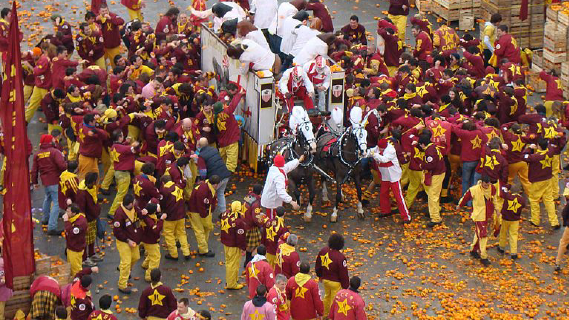 Zdjęcie przedstawia bitwę na pomarańcze. W centrum kadru widać wóz zaprzężony w konia. Wokół jest tłum ludzi, głównie mężczyzn. Na ulicy leżą pomarańcze. Nie które z osób mają owoce w rękach. Po prawej stronie stoją skrzynie służące do transportu pomarańczy.
