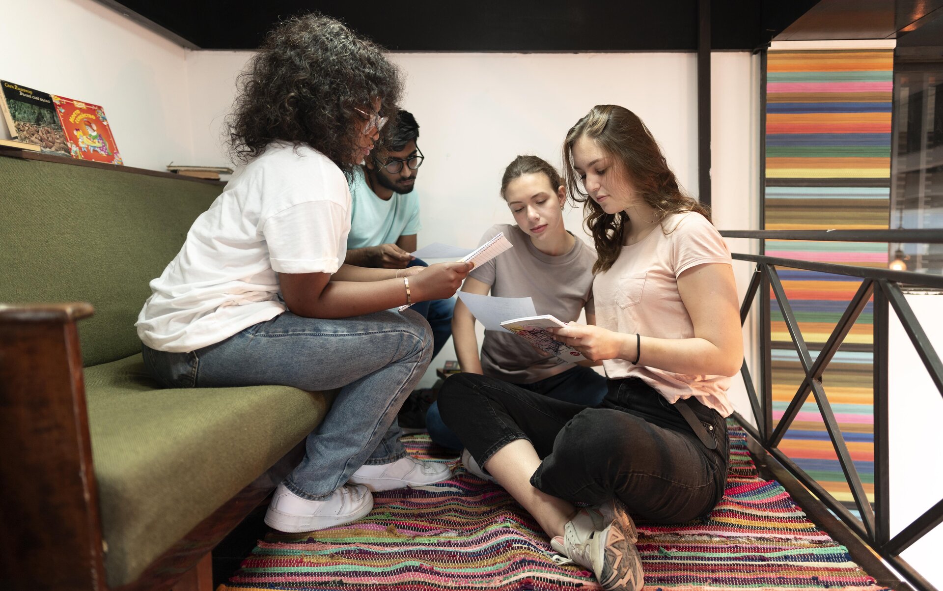 Zdjęcie przedstawia cztery osoby skupione nad wspólnym zadaniem. Dziewczyna i chłopak siedzą na kanapie a obok na podłodze siedzą po turecku dwie nastolatki. Osoby na kanapie mają ciemną skórę i ciemne włosy, dziewczyny siedzące na podłodze mają jasną skórę i ciemno blond włosy. Transkrypcja nagrań znajduje się poniżej.