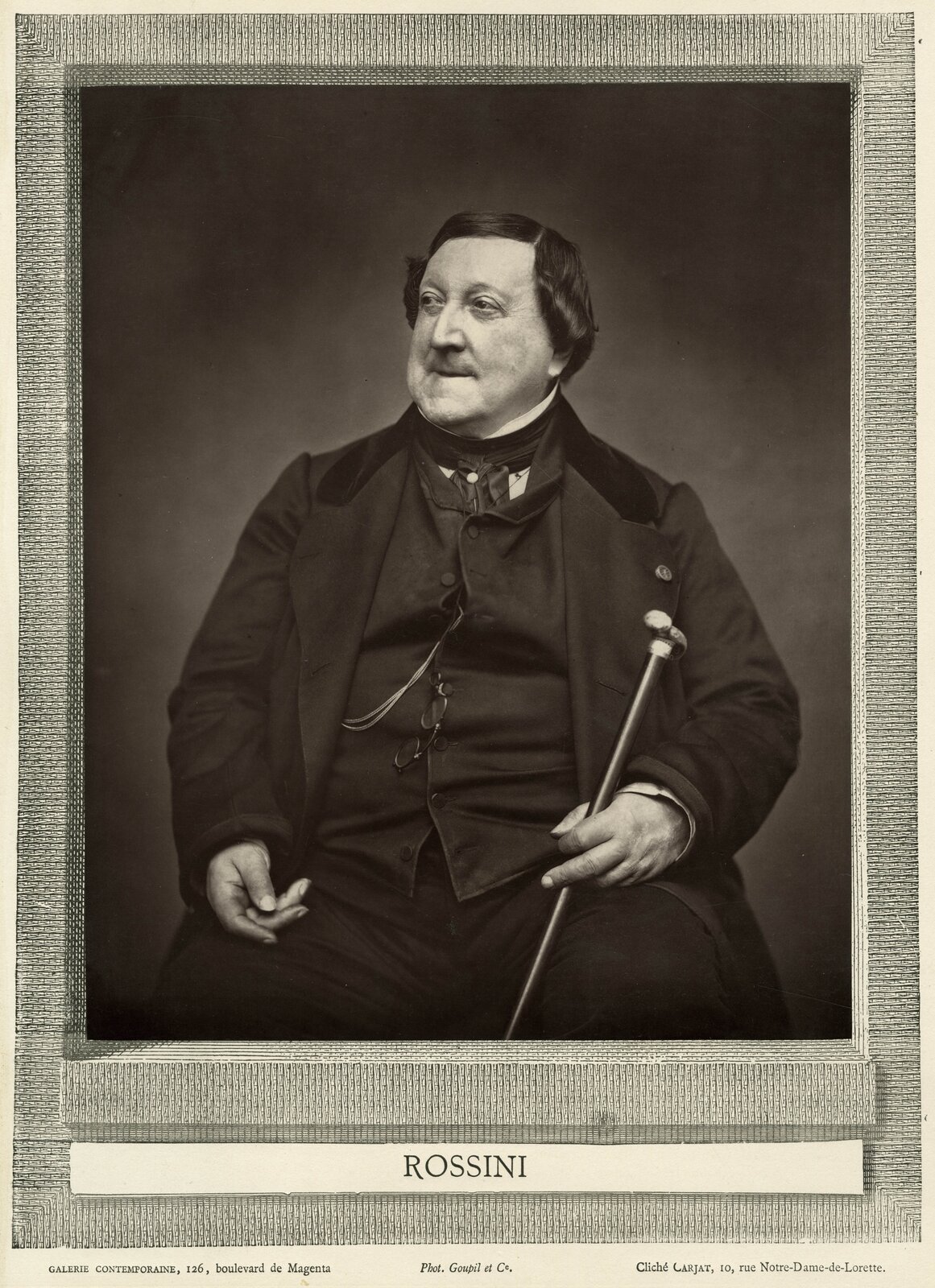 Ilustracja przedstawia portret Gioacchino Rossiniego nieznanego autora. Rossini pozuje siedząc. W dłoni trzyma laskę, a na torsie zawieszone ma okulary.