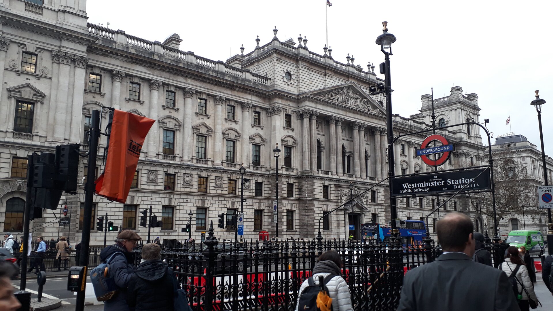Fotografia monumentalnego, ogromnego pałacu. Na fasadzie ozdobne kolumny, duże okna. Przed pałacem ulica, wejście do metra.