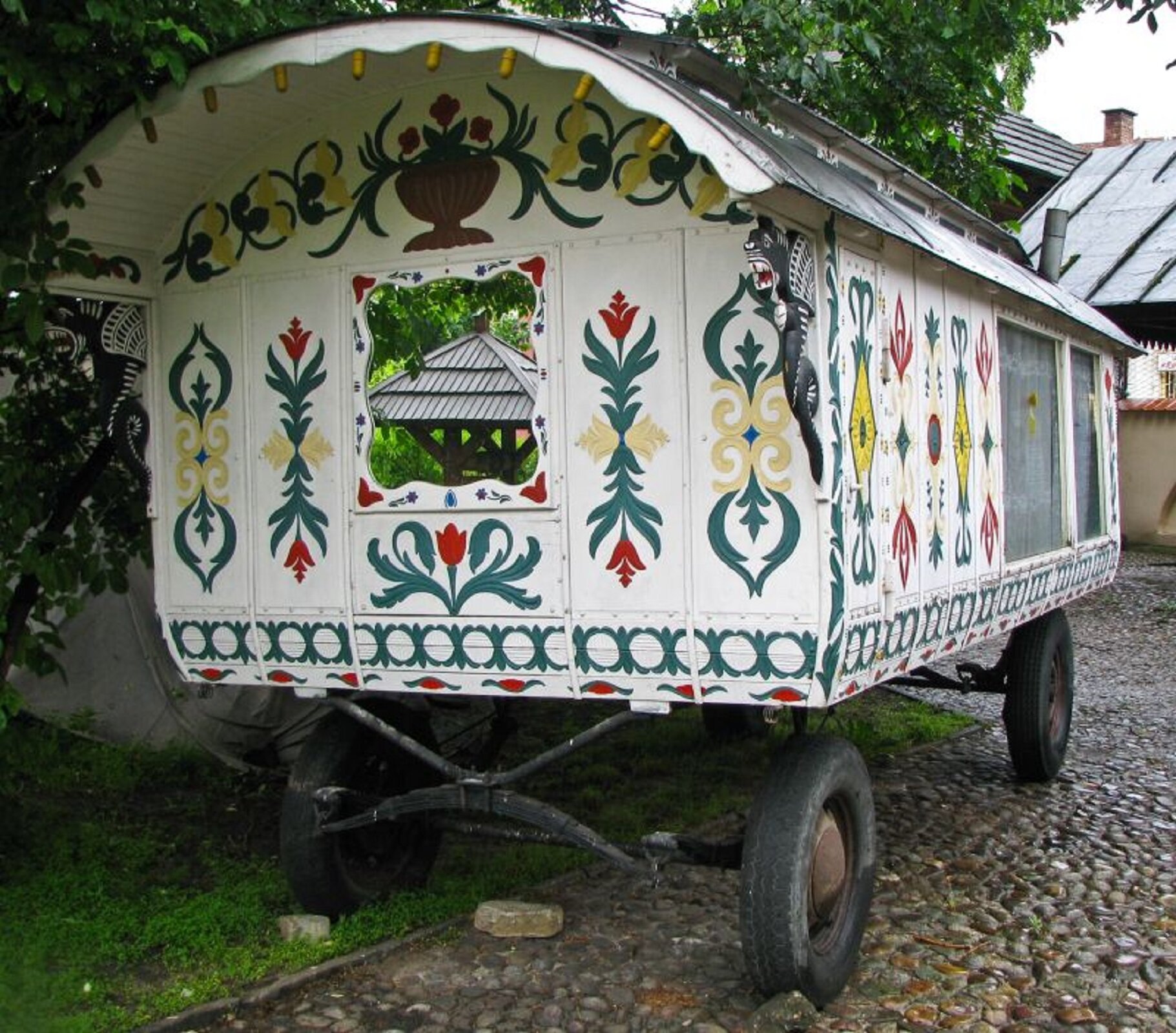 Ilustracja przedstawia bogato zdobiony wóz romski. Wóz pomalowany jest z zewnątrz na biało, umieszczone są pod dachem ozdobne gryfy i smoki z drewna.