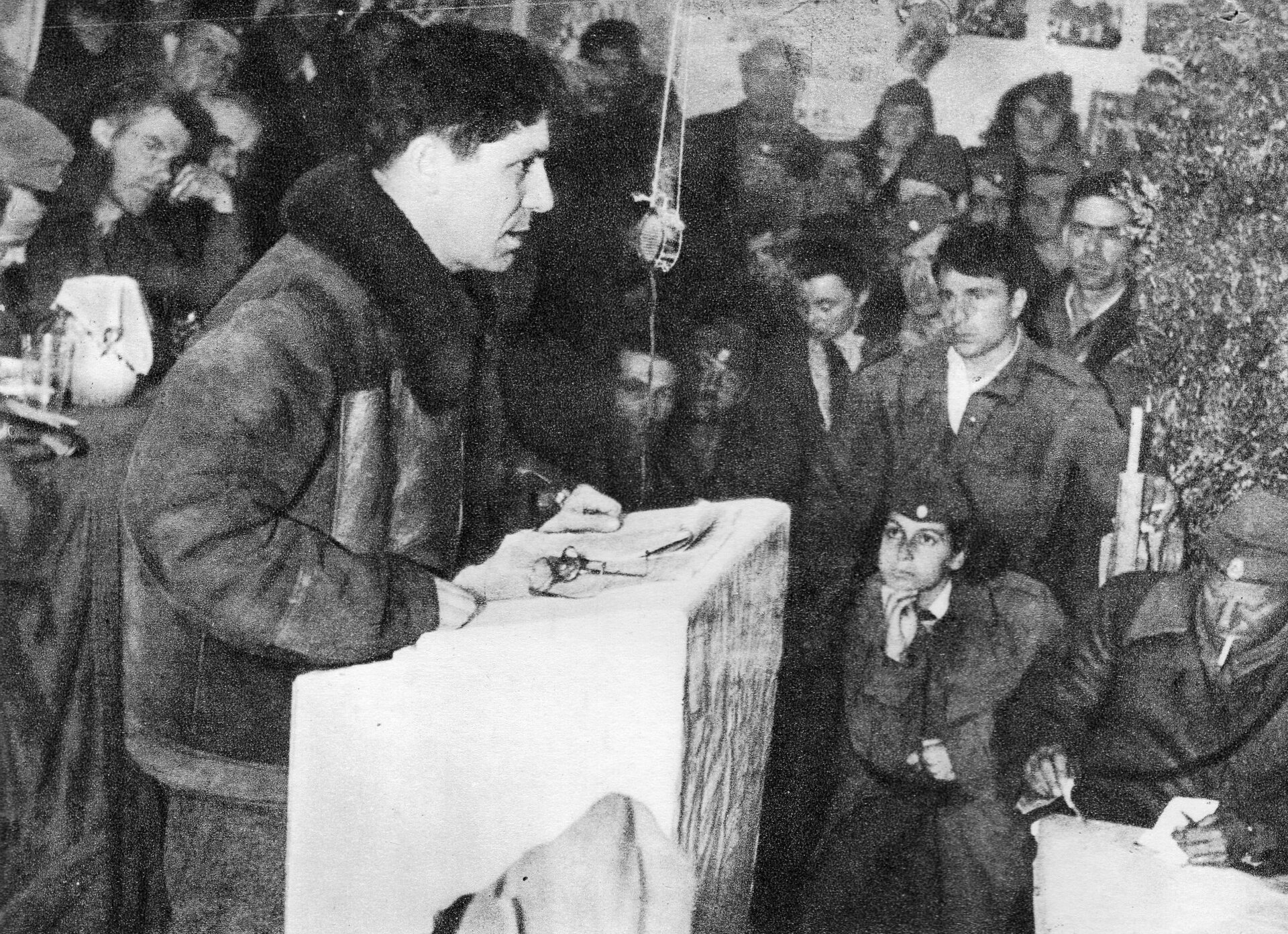 Czarno0białe zdjęcie przedstawia mężczyznę przemawiającego z białej mównicy do tłumu słuchaczy. Jest on ubrany w kurtkę. Ma krótkie ciemne włosy. 