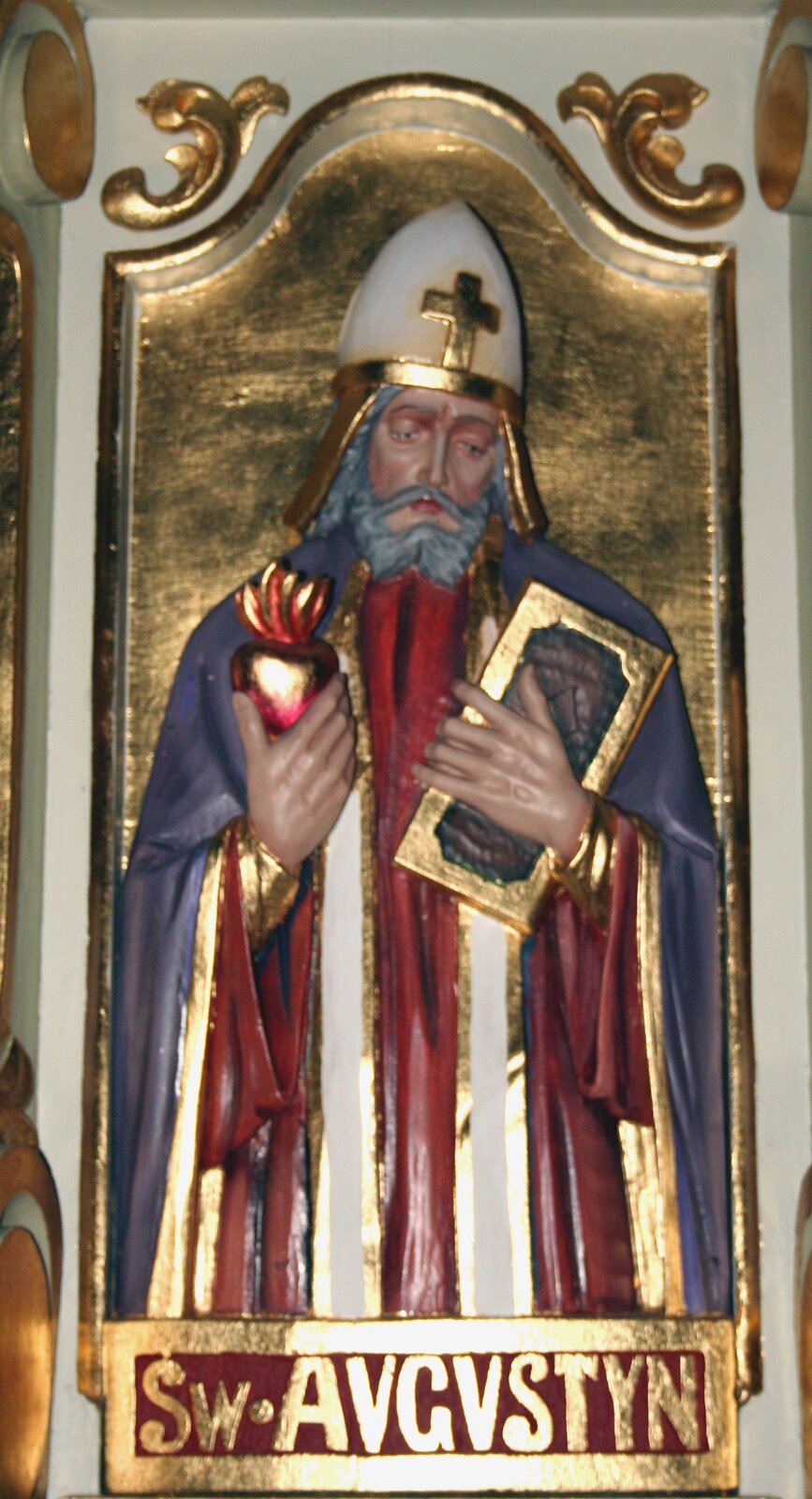 Zdjęcie przedstawia płaskorzeźbę, na której uwieczniony jest święty Augustyn. Mężczyzna ubrany jest w szaty biskupie. Na głowie ma tiarę. W prawej ręce trzyma serce, w lewej księgę.