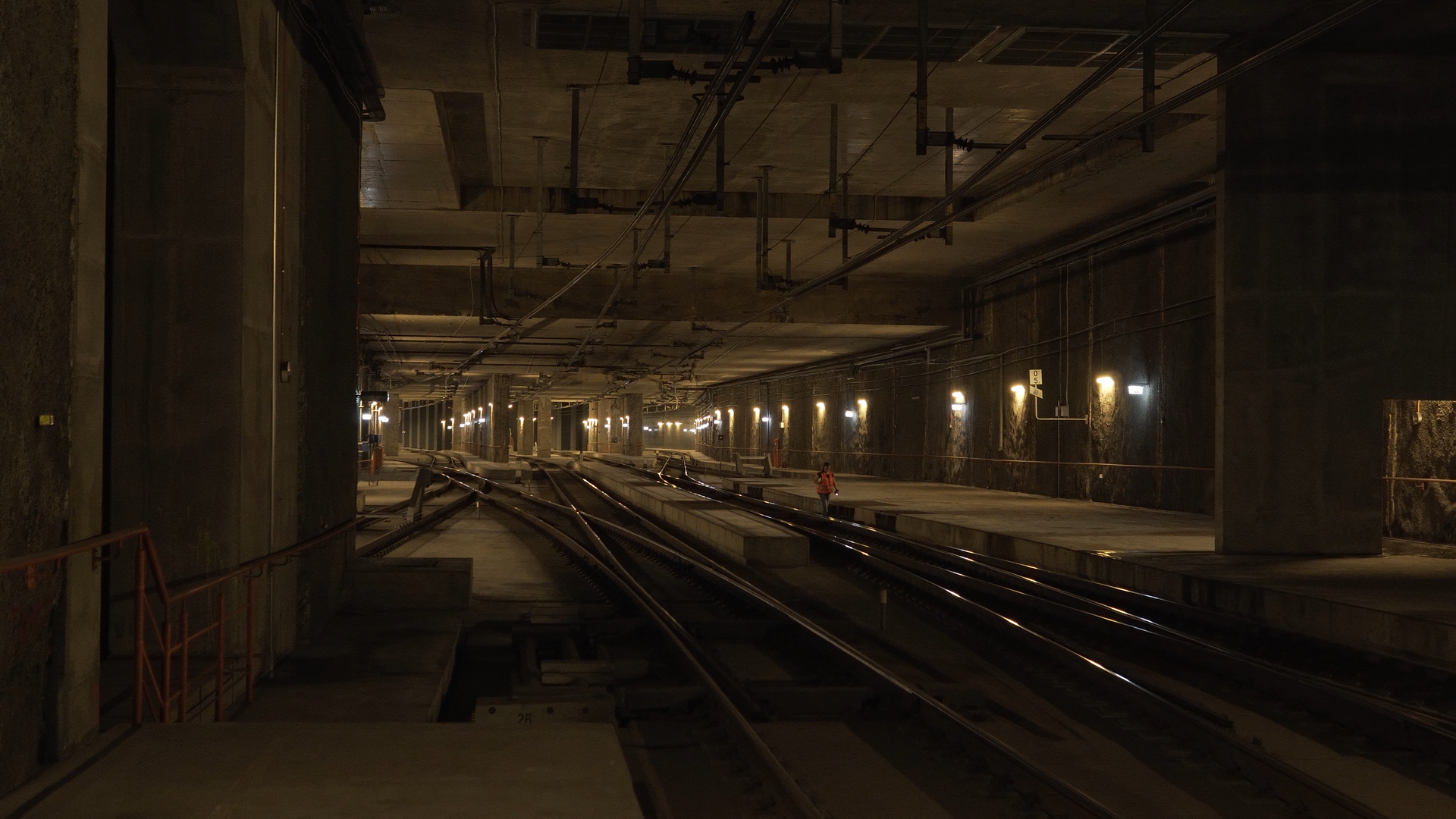 Ilustracja ukazuje tunel kolejowy wykonany z betonu, którego wnętrze jest jasne i dobrze oświetlone. Na ścianach tunelu umieszczone są urządzenia oświetleniowe, które zapewniają odpowiednią widoczność. W tle wyróżnia się dwutorowe torowisko oraz pracownicy kolei noszący odblaskowe kamizelki, co sugeruje, że wykonują prace na linii kolejowej. Obok torowiska przebiegają chodniki, których używają pracownicy. Na suficie tunelu można dostrzec wiszące metalowe elementy, dodając tym samym industrialnego charakteru całej konstrukcji.