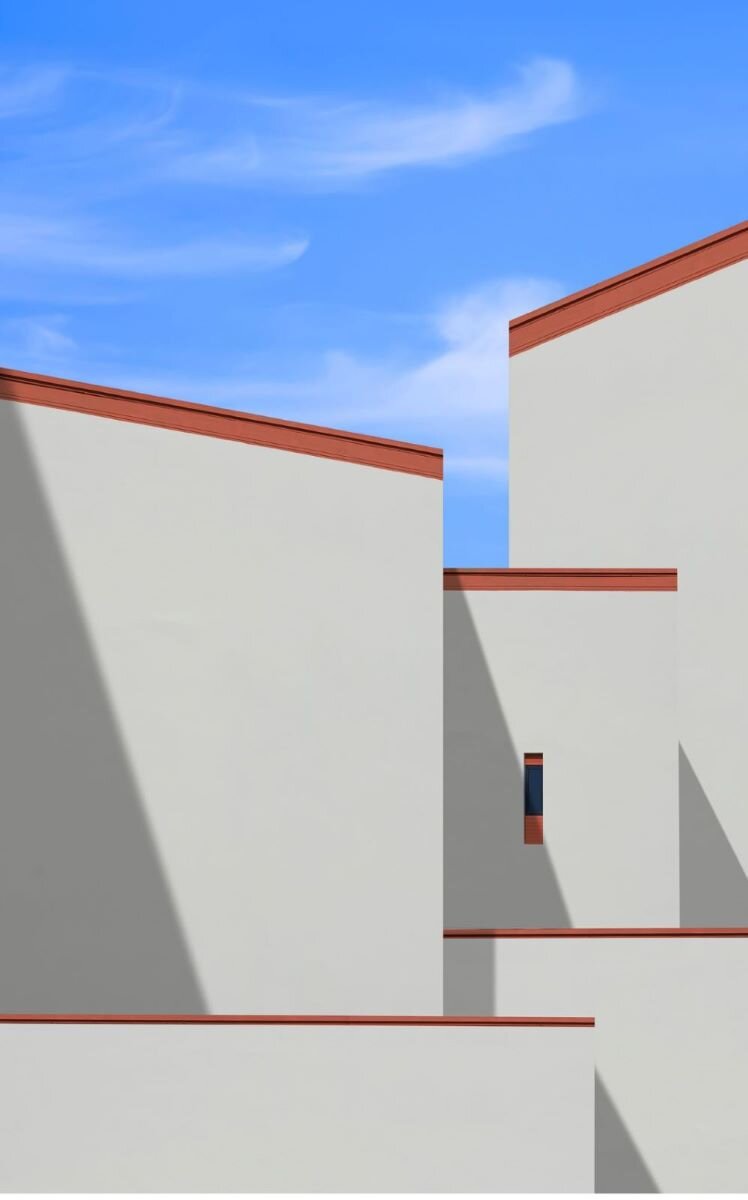 Ilustracja przedstawia zbliżenie na fragmenty elewacji obiektów w zwartej zabudowie. Są to zewnętrzne ściany o różnej wysokości, zachodzące jedne na drugie. Każda z nich jest w jednakowej kolorystyce: białe ściany zwieńczone wąskimi czerwonymi pasami. 