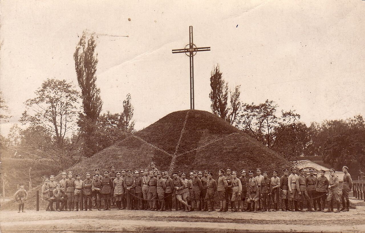 Zdjęcie przedstawia niewielki kopiec, na szczycie którego stoi duży krzyż. U stóp kopca stoi duża grupa mężczyzn w mundurach. Pozują do zdjęcia.